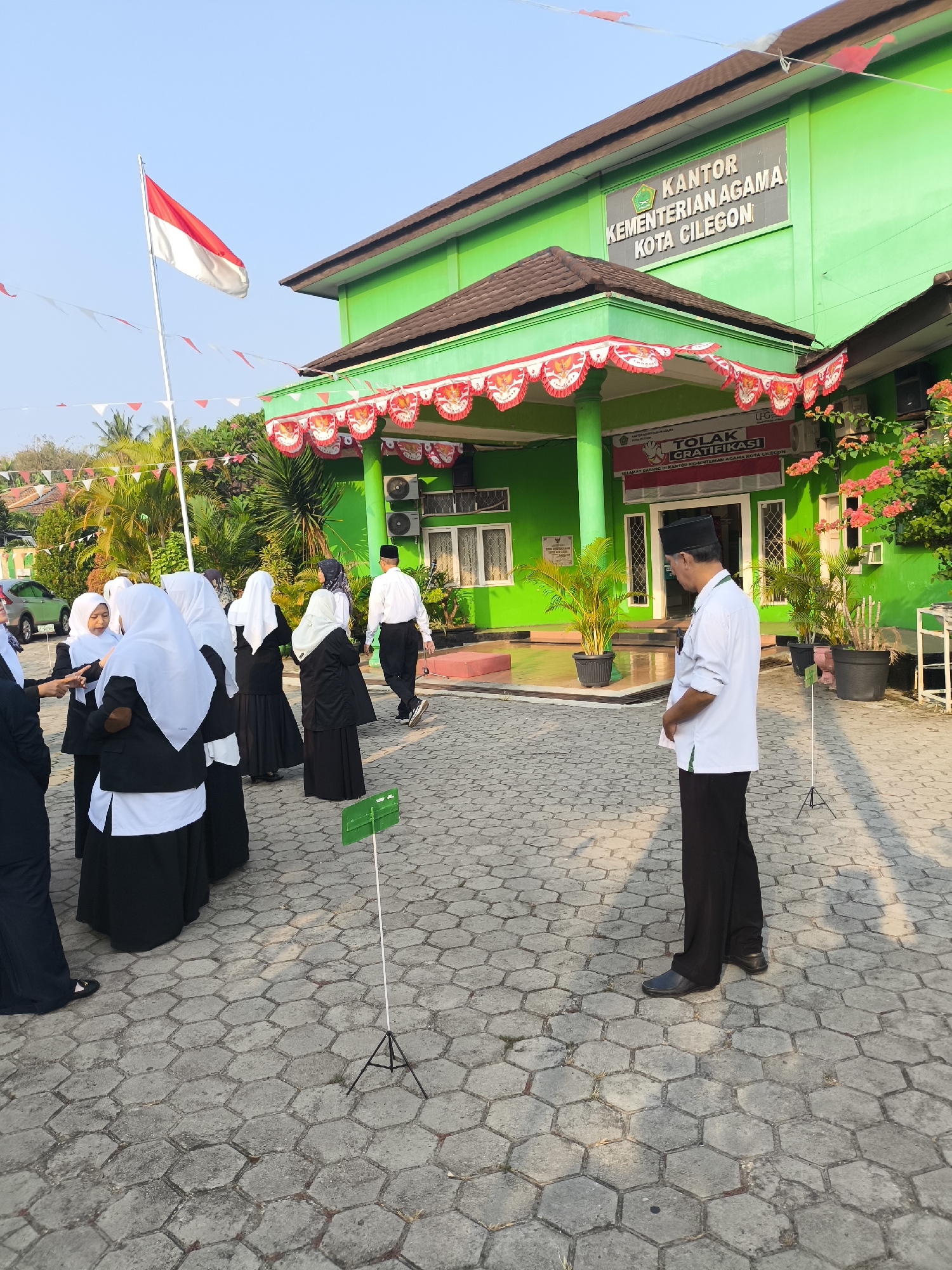 The image depicts the exterior of the "KANTOR KEMENTERIAN AGAMA KOTA CILEGON" (Ministry of Religious Affairs Office, Cilegon City) in Cilegon, Indonesia. The building is painted bright green with a brown tiled roof and is adorned with red and white festive banners. An Indonesian flag flies from a flagpole to the left of the building. Several people are gathered in front of the building, including a group of women in black and white hijabs and a man in a white shirt and black pants wearing a peci cap. The ground is paved with hexagonal grey bricks. A green sign on a tripod is visible in the foreground. The overall atmosphere appears to be a formal gathering or event.