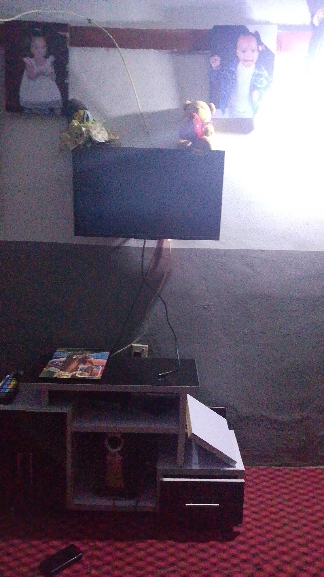 In an indoor scene with a red patterned carpet, a dark gray wall, and a light-colored ceiling, a flat-screen television is mounted on the wall, displaying a dark, blank screen with a faint orange light at the bottom edge. Below the television, a dark brown media console features a black top with a magazine or book placed on it. The console has multiple shelves and compartments; one open compartment on the left contains a yellow object, and the right side of the console has a white rectangular object leaning against it.
On the wall above the television, two framed pictures are visible. The left frame contains an image of a young child in a white dress. To the right of this, a teddy bear toy is placed on the wall shelf, holding a red heart. To the far right, another framed picture shows a young child with dark hair and a light-colored jacket.
Various cables are visible hanging from the television and plugged into an electrical outlet on the wall. A remote control is on the left side of the media console, and a dark rectangular object, possibly a phone, lies on the red carpet in the foreground. The lighting in the scene is dim, with a bright light source emanating from the upper right portion of the image, suggesting a window or strong artificial light.