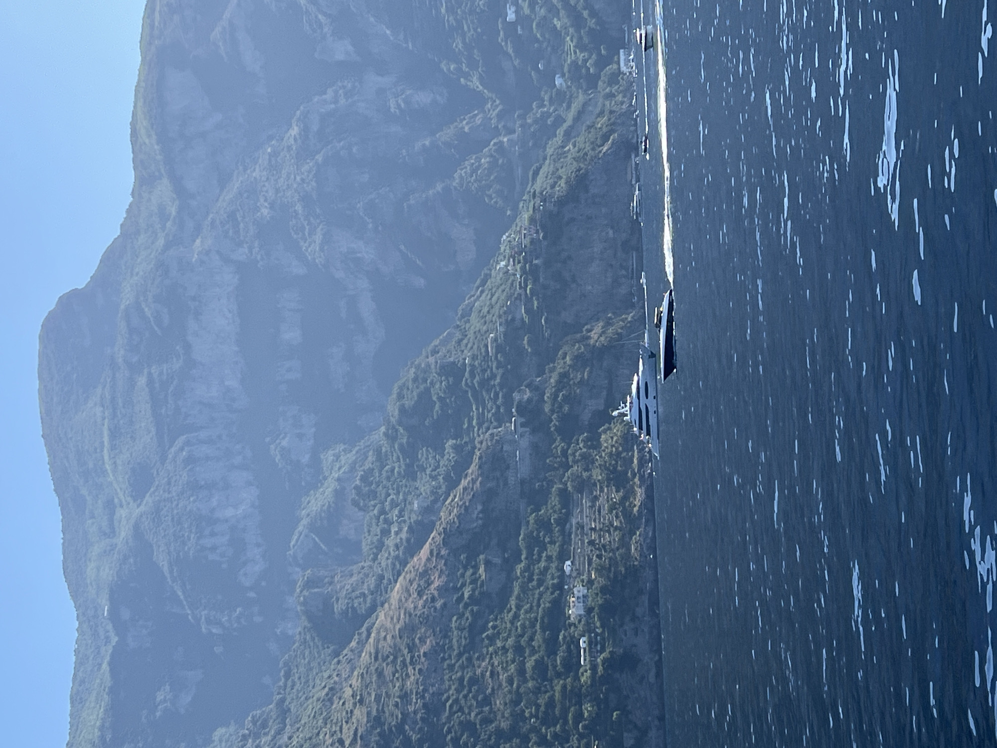 The image depicts a scenic view of the Amalfi Coast near Arienzo, Italy. In the foreground, the deep blue water of the Tyrrhenian Sea is visible, with gentle waves and reflections of light. Several boats are navigating the water; a large white yacht is prominent on the left, followed by a smaller dark-colored speedboat leaving a white wake. In the background, steep, verdant mountains rise dramatically from the sea. The mountains are covered in dense vegetation and dotted with small buildings, indicative of a coastal settlement. The terrain appears terraced, suggesting agricultural use of the slopes. The sky above is a clear, bright blue. The overall impression is one of a beautiful, sun-drenched day on the Italian coast.