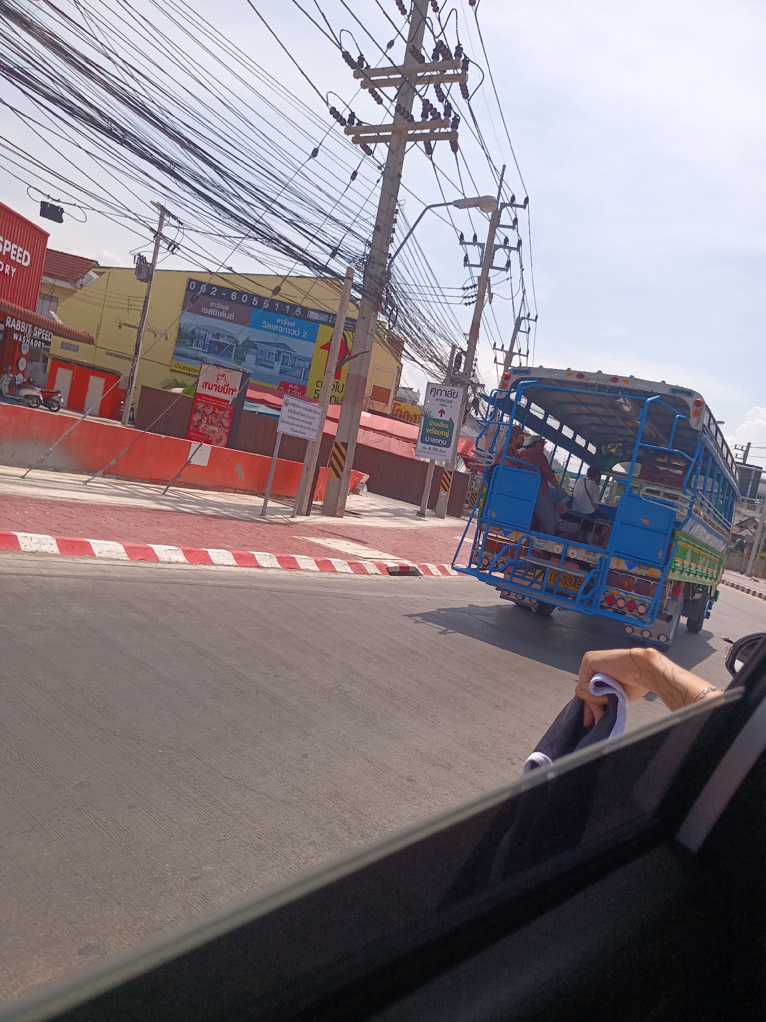 The image shows a view from inside a car, looking out at a street in Hua Hin City Municipality, Thailand. On the road, a blue Songthaew (public transport vehicle) is moving, occupied by people. Sidewalks with red and white striped edges are visible, along with buildings that display Thai script and advertisements. Overhanging wires are abundant, supported by utility poles. The image captures a typical street scene with local transport and infrastructure.