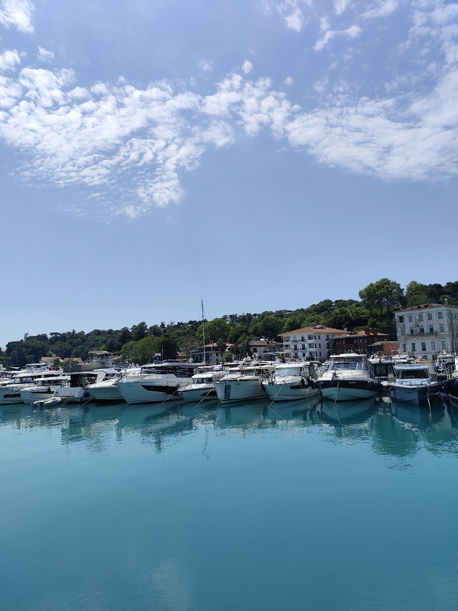 This image captures a serene daytime view of a marina in Sarıyer, Turkey. The scene is dominated by a tranquil body of turquoise water in the foreground, reflecting the bright sky and the numerous boats docked along the shore.
A long line of private yachts and motorboats, predominantly white and light-colored, are moored side-by-side. These vessels vary in size and design, featuring clean hulls and intricate details. Visible text on some boat hulls includes "A.SY.R" (partially obscured) on a boat to the left, "NEZDEMAT" on a prominent boat in the center-right, and "A.2241" (partially obscured) on another. Mooring lines connect the boats to the dock, although the dock itself is not clearly visible.
Behind the marina, a lush, green hillside rises, densely covered with trees and verdant foliage. Several buildings are nestled among the trees, including residential structures and a large, distinct white building with multiple windows and balconies on the right side of the frame, showcasing local architecture.
The sky above is a clear, bright blue, accented by scattered patches of fluffy white clouds that stretch across the upper portion of the frame. The strong, even lighting suggests it is midday or early afternoon, indicative of pleasant, sunny weather with calm conditions. There are no people visible in the image, emphasizing the peaceful and quiet ambiance of the marina. The overall impression is one of a picturesque coastal retreat.