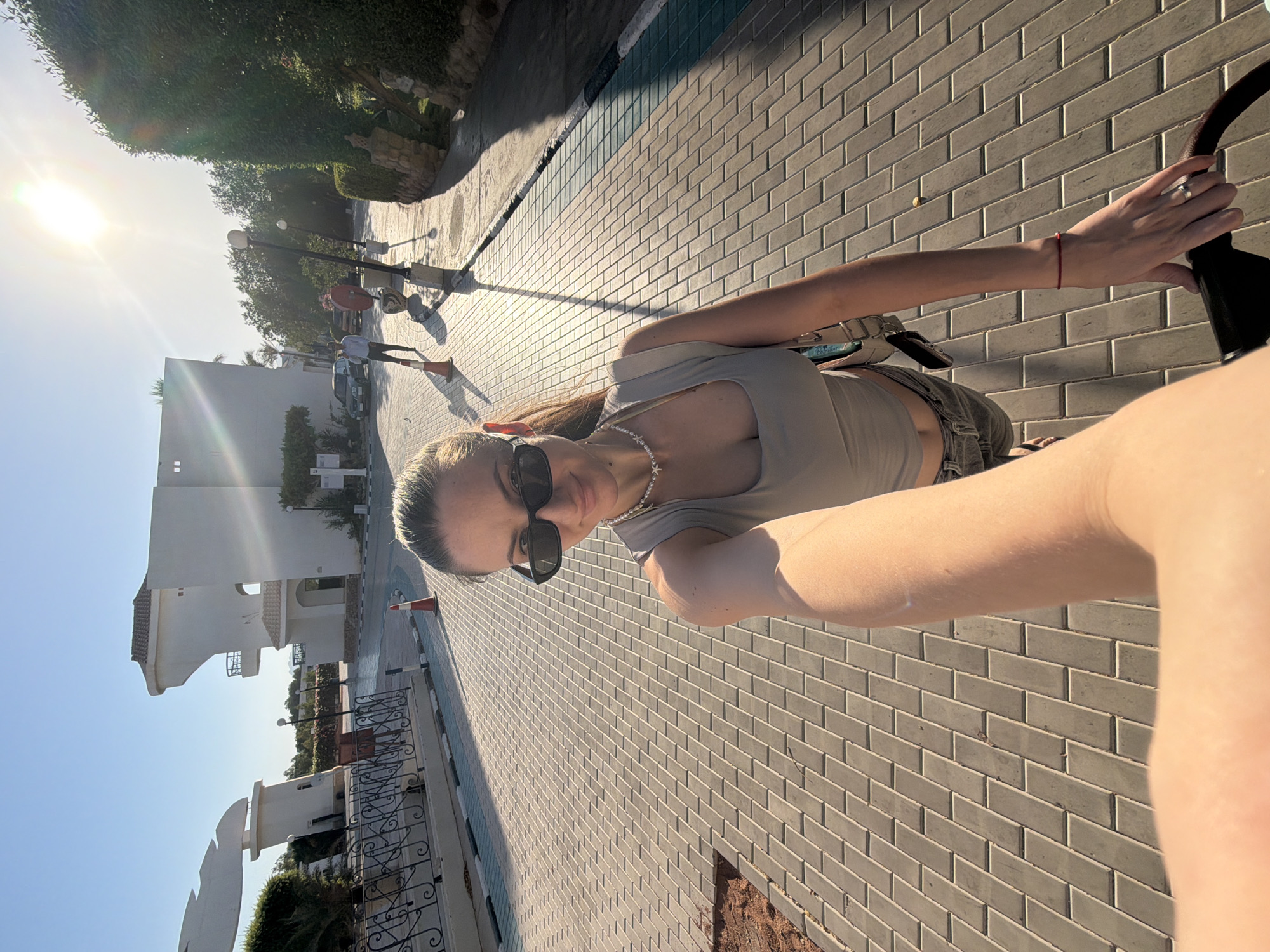 A woman in sunglasses and a grey top is taking a selfie on a paved street. The sun is shining brightly in the sky. In the background, there are buildings, trees, and a few people. The scene appears to be in a resort or residential area in Sharm El Sheikh, Egypt, given the architectural style and the presence of palm trees. The woman is wearing a pearl necklace and a red bracelet on her left wrist. She is also holding onto a stroller or some similar wheeled object. The pavement is made of rectangular grey bricks.