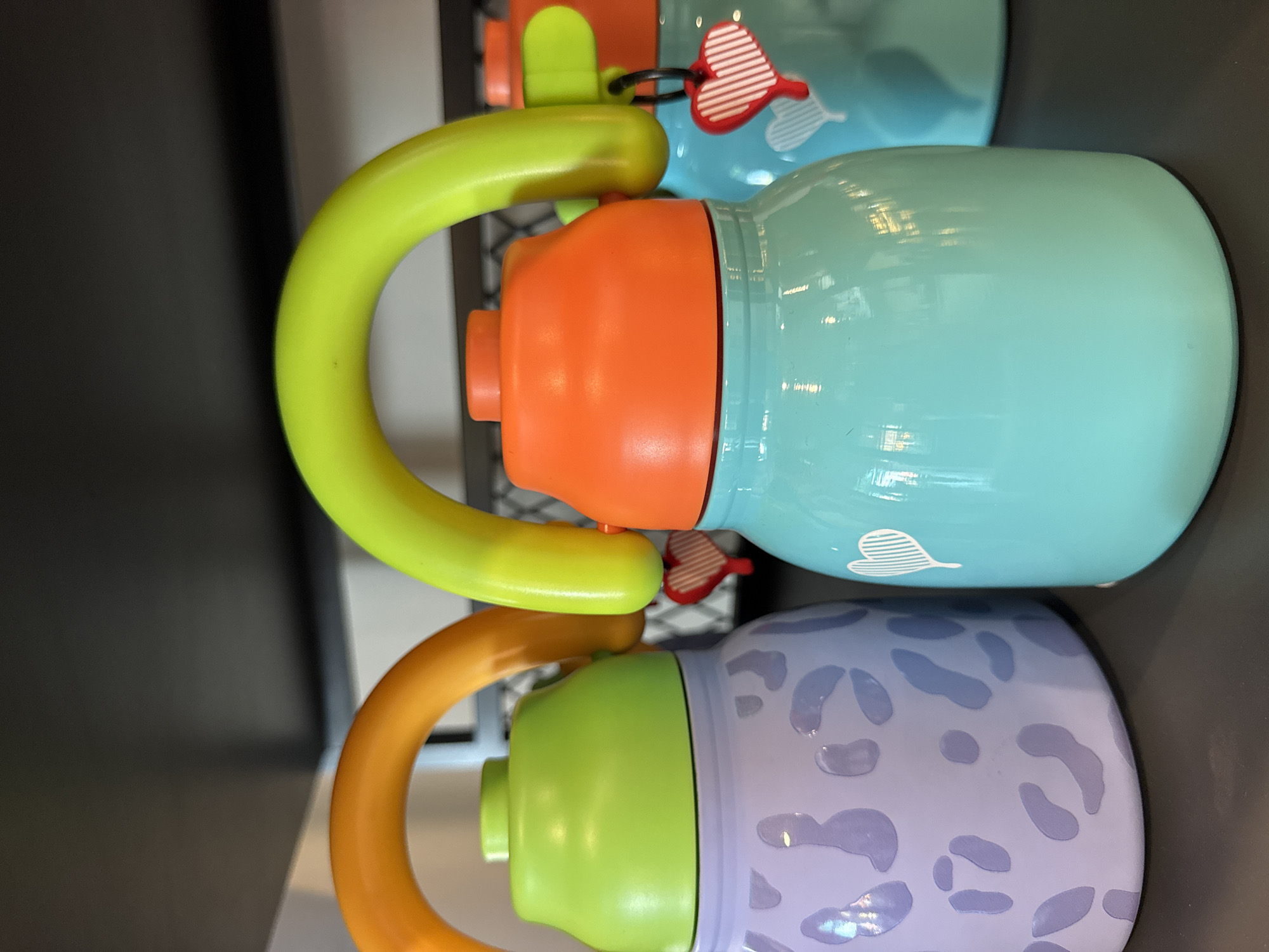 The image features two vibrantly colored water bottles or thermoses resting horizontally on a dark surface. The upper bottle has a smooth, light teal body, a bright orange cap assembly, and a prominent, C-shaped lime green handle. A decorative red and white striped heart charm, attached via a black ring, hangs from the bottle's top. A subtle white heart design is also visible on the teal body.
Below it, the second bottle showcases a light purple body patterned with darker purple, irregular leopard-spot-like designs. This bottle is fitted with a light green cap and a large, C-shaped orange handle. Both bottles exhibit a glossy finish, reflecting ambient light. The background is dark and out of focus, suggesting an indoor setting, possibly a retail display or shelf. There is no activity depicted, and no people are present. No text is visible on any of the objects. The time of day and weather cannot be determined from the image.