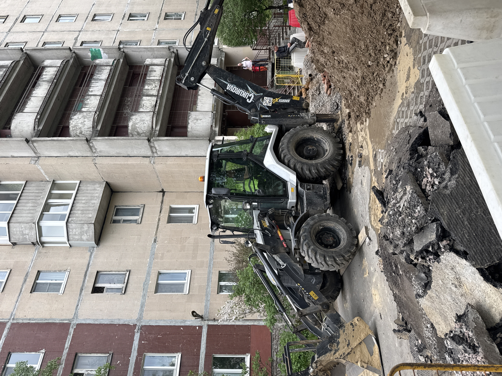 The image shows a backhoe excavator working near a building. The excavator, a "Cukurova BGS 888," is positioned on a damaged asphalt surface, with broken pieces scattered around. A pile of dirt is adjacent to the excavation. Two people are on the steps of the building, overlooking the work. The building itself is multi-storied with tiled exterior walls and cantilevered balconies. The scene is typical of construction or road repair work in an urban environment. Based on the architecture of the building, and the make of the backhoe, it is highly probable that the location of this image is the city of Saint Petersburg, Russia.