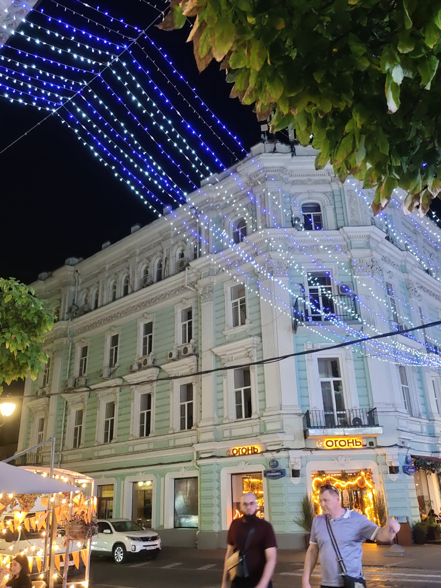This is a nighttime, slightly tilted, eye-level, medium shot of a building in Rostov-on-Don, Russia, decorated with numerous blue and white string lights. The building appears to be an older, ornate structure with light green siding and white architectural details. Several windows are visible, some with air conditioning units mounted below them. The building's corner has a sign with orange lettering that reads "ОГОНЬ", which translates to "FIRE" in English. Two men are walking in the foreground. The man on the left is wearing a dark burgundy shirt and the man on the right is wearing a light blue polo shirt. Both men are carrying bags. To the left of the building, part of an outdoor cafe or market stall is visible with string lights and orange banners. A white car is parked on the street in front of the building. Above the building, dark foliage from a tree hangs into the frame, with some leaves appearing dry or brown. The sky is dark and appears to be clear.