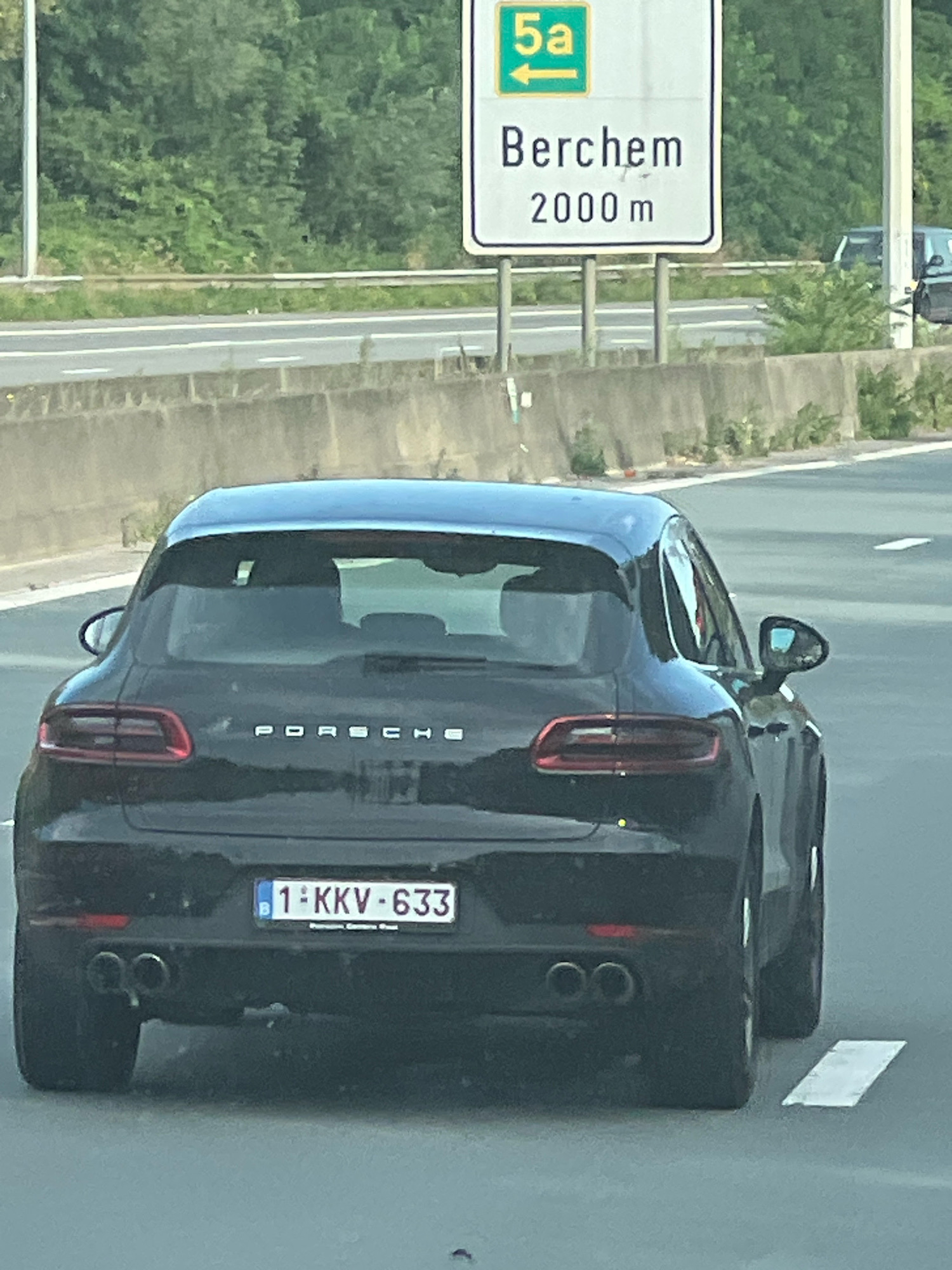 The image shows the rear of a black Porsche Macan driving on a highway. A road sign indicates "Berchem 2000 m" and "5a" with an arrow pointing left. The car has a license plate that reads "1-KKV-633". The environment appears to be a suburban highway with trees and vegetation visible in the background. No people are clearly visible. The highway has concrete barriers and road markings. The image is taken from a slightly elevated perspective, looking down at the car. The location is Edegem, Belgium, as indicated by the road sign referencing Berchem, which is a district of Antwerp and close to Edegem.
