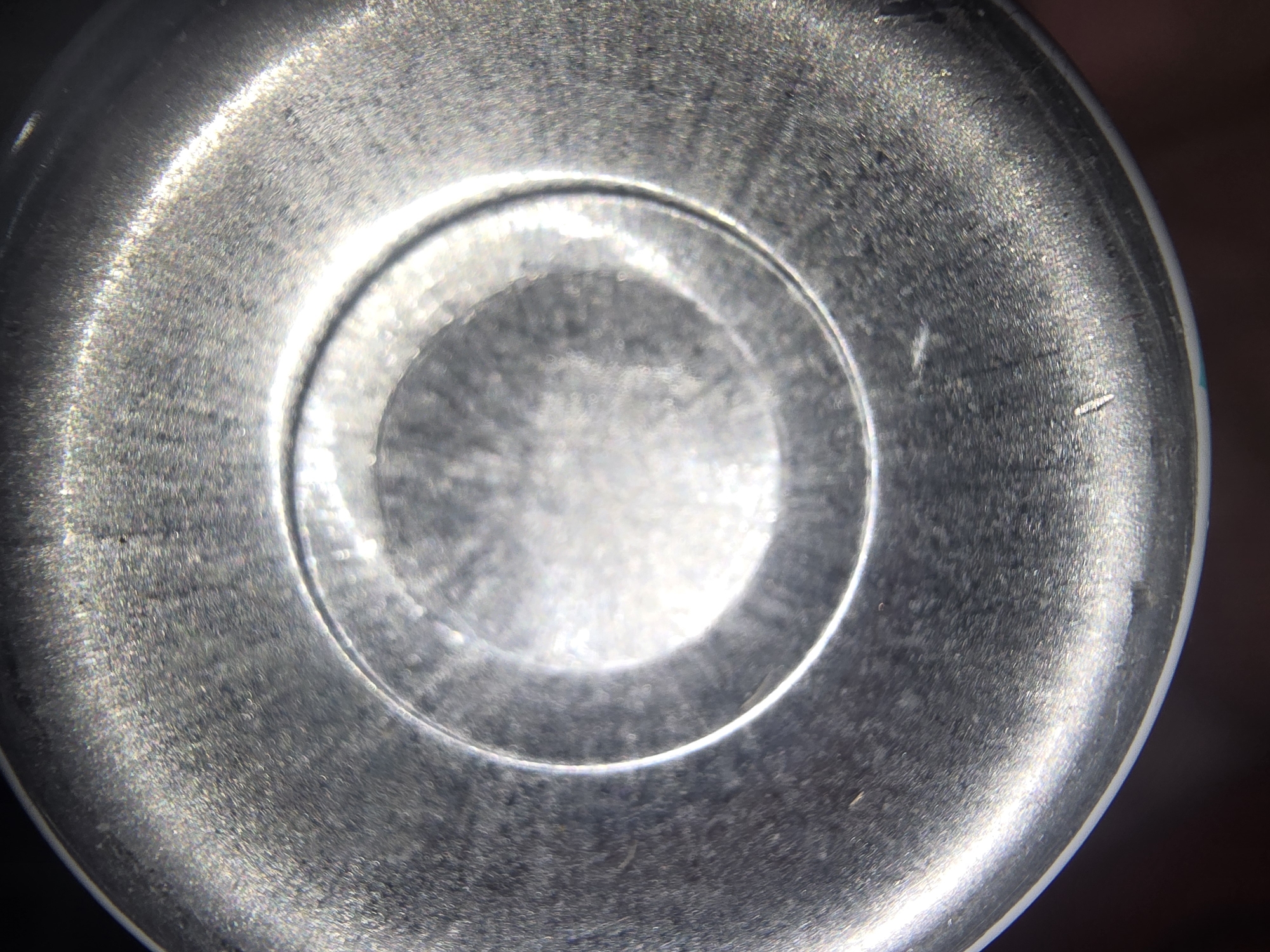 The image is a close-up, macro photograph of the bottom of a metal can. Concentric circles are visible, indicating the manufacturing process. The surface is textured and shows signs of wear and possibly some dust or residue. The lighting creates a metallic sheen with highlights and shadows. The background is dark and out of focus. There are no people, animals, or other objects visible. The image is abstract in nature and does not depict a specific location in Ulan-Ude, Russia, or any other geographical area.