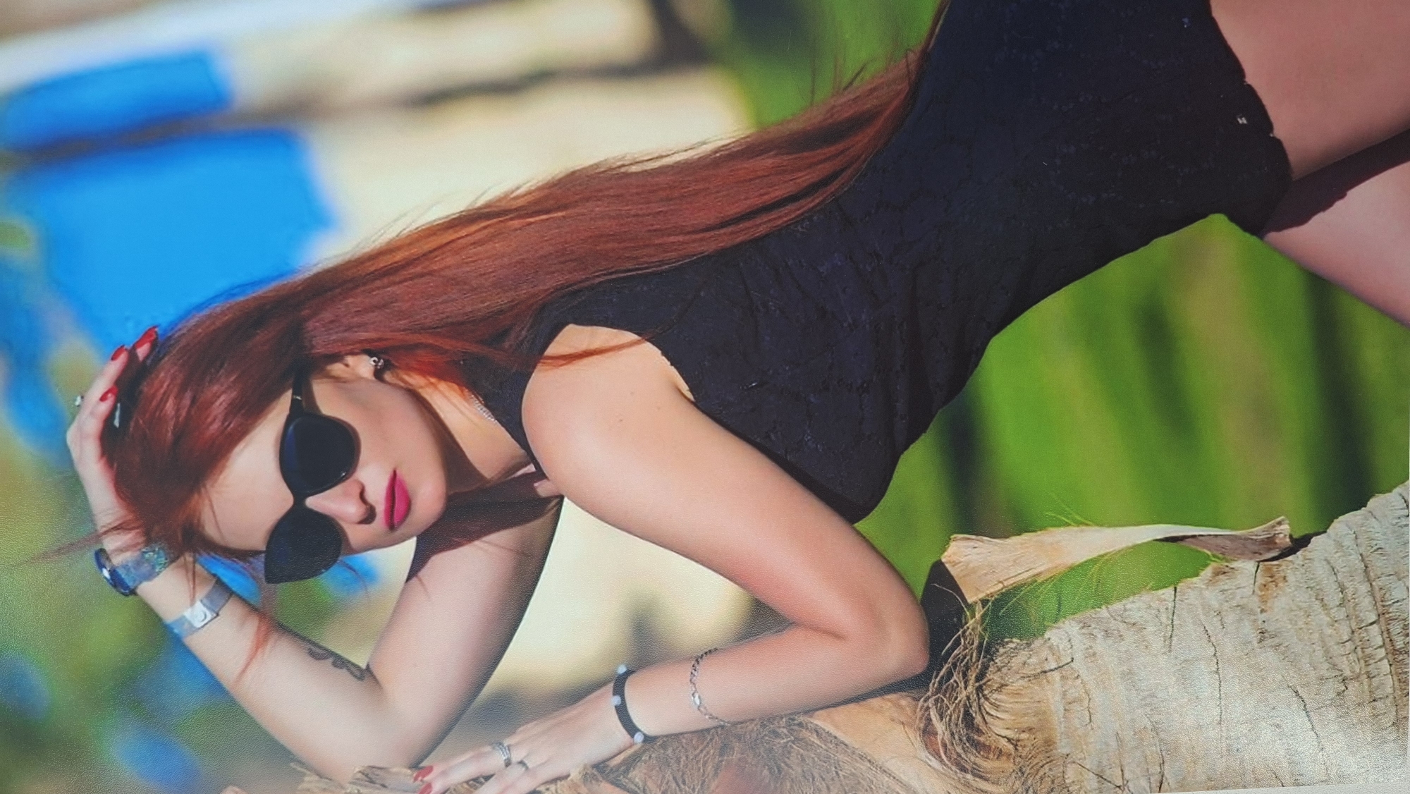 A woman with long red hair, sunglasses, and a black dress lies on a textured surface, possibly wood. She is adorned with wrist jewelry and has red nail polish. The background is a blur of green and blue, possibly suggesting an outdoor location.
