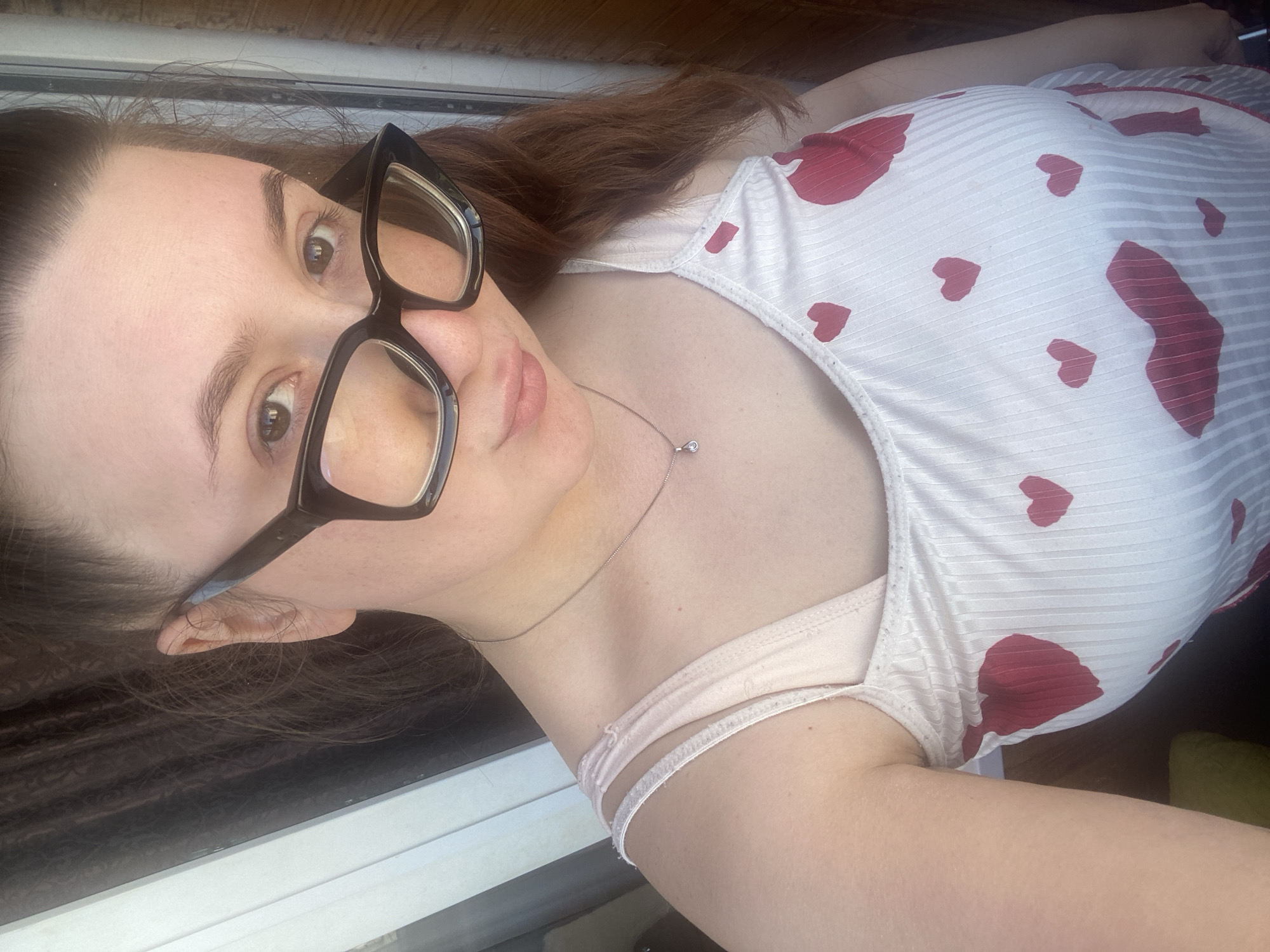 A woman with brown hair and wearing large black-rimmed glasses looks directly at the camera. She is wearing a white tank top with red heart-shaped designs. A delicate silver necklace adorns her neck. The background appears to be a wooden surface. The location is not discernible from the image and cannot be confirmed as Chaikovsky, Russia.