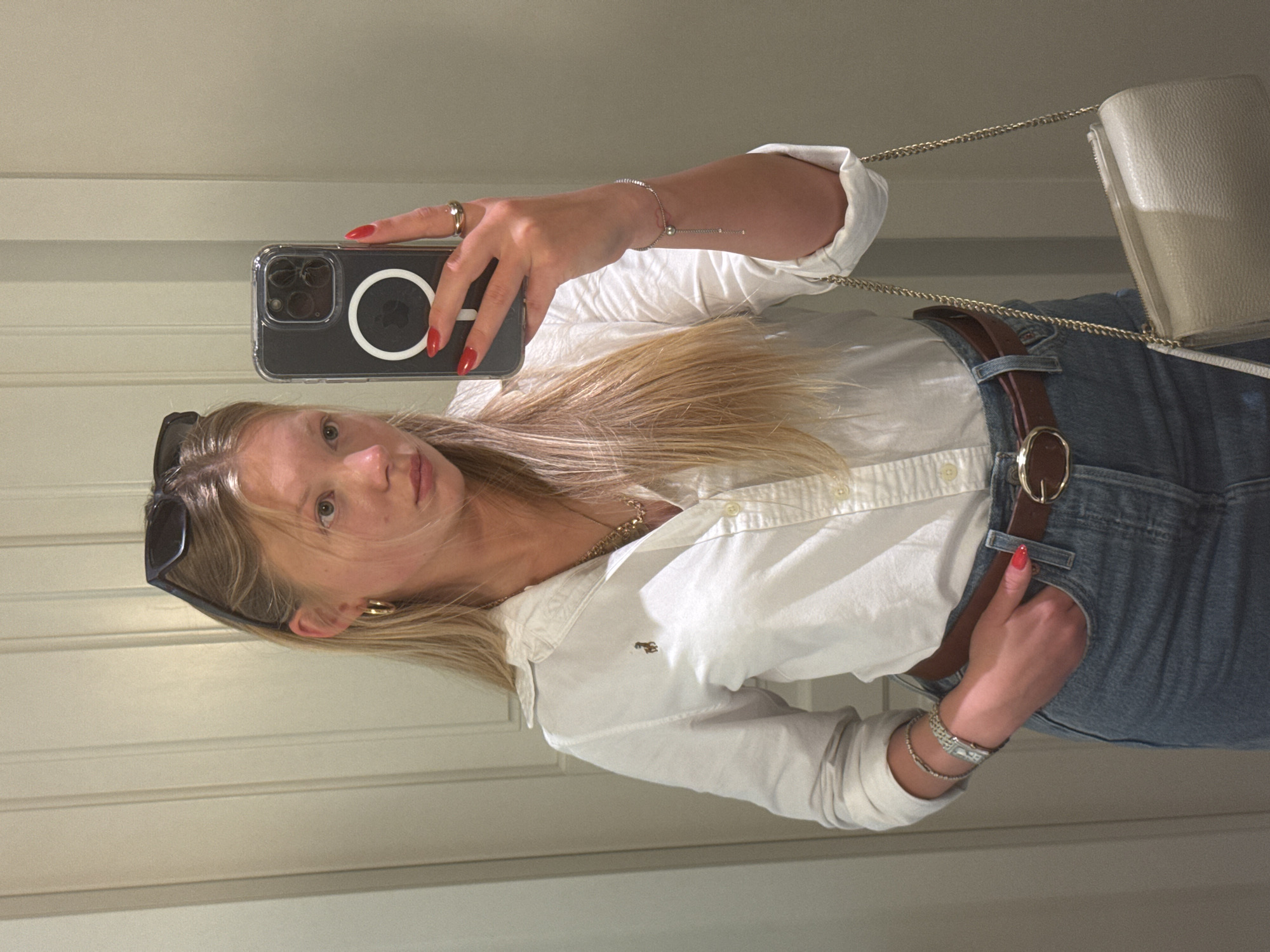 A woman takes a selfie in what appears to be a dressing room or closet. She holds an iPhone in her right hand, the camera facing her, and a beige-colored purse with a chain strap hangs at her side. The woman has blonde hair and is wearing sunglasses perched on her head, gold hoop earrings, a gold necklace, a white button-down shirt, a brown belt, and blue jeans. Red nail polish is visible on her fingers. She is looking at the camera. The background consists of vertical white panels. The location of the image is unknown.