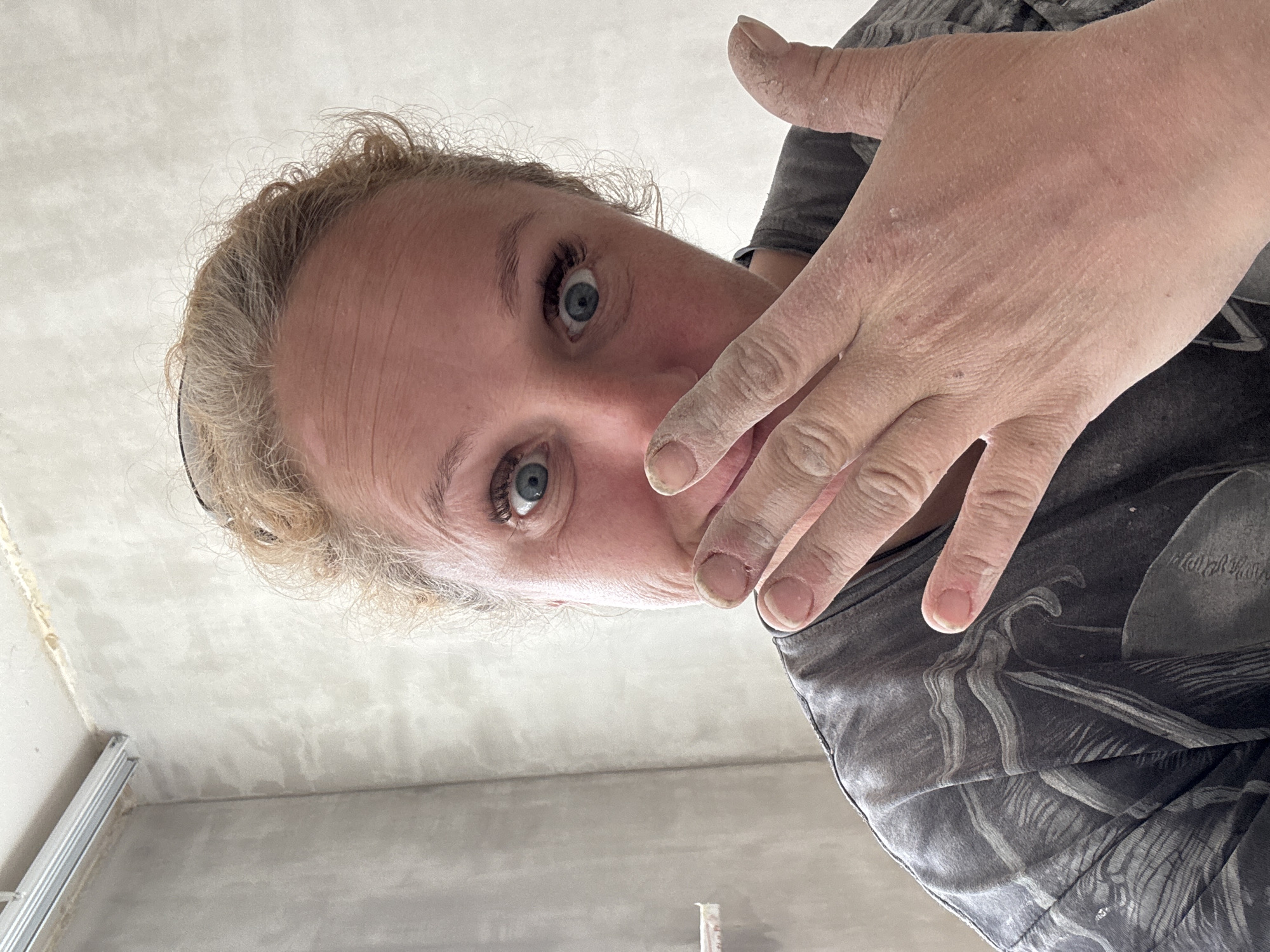 A low-angle, close-up shot shows a person's face and hand. The person has fair skin, blue eyes, and curly blond hair. Their hand is positioned in front of their face, with the fingers slightly curved. The hand and face appear to have dust or powder on them. The person is wearing a gray patterned shirt. The background includes a corner of a room with grey walls and a metal frame.
There is no basis in the image to conclude that the location is Chaikovsky, Russia.