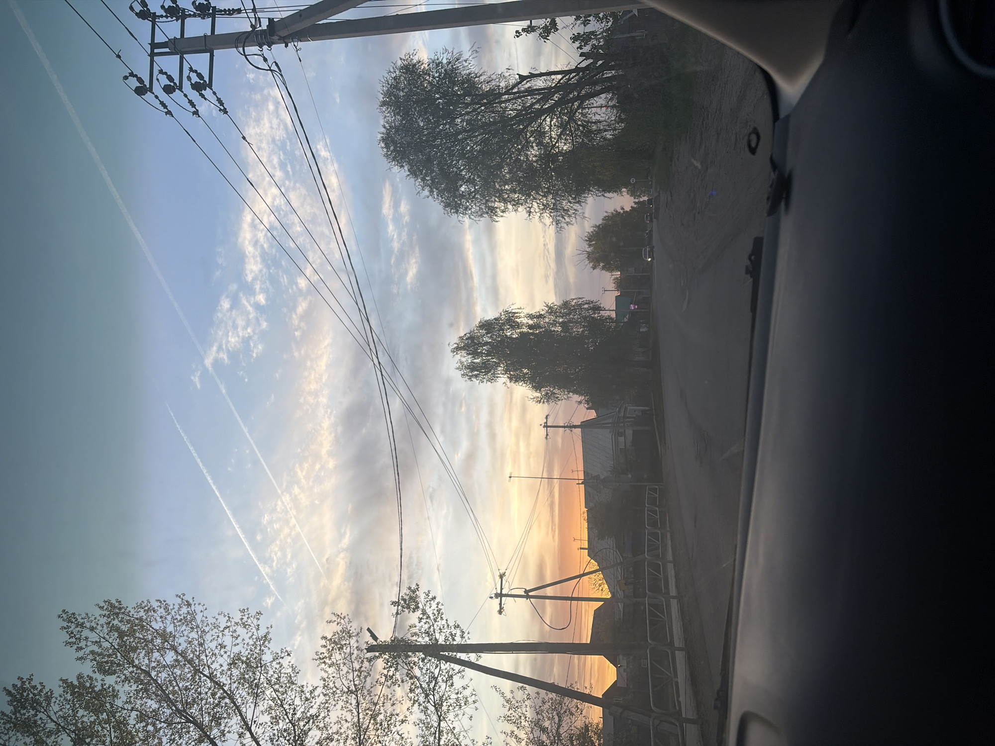 The image depicts a view from inside a vehicle, likely a car, looking out onto a street in Chaikovsky, Russia. The sky shows a sunset with orange and yellow hues blending with light blue and white clouds. Power lines crisscross the sky, attached to utility poles on either side of the road. Trees line the street, some silhouetted against the sunset. A road stretches into the distance with buildings and fencing visible along the roadside. The inside of the vehicle is visible in the foreground, showing the dashboard and part of the door. There are contrails visible in the sky.