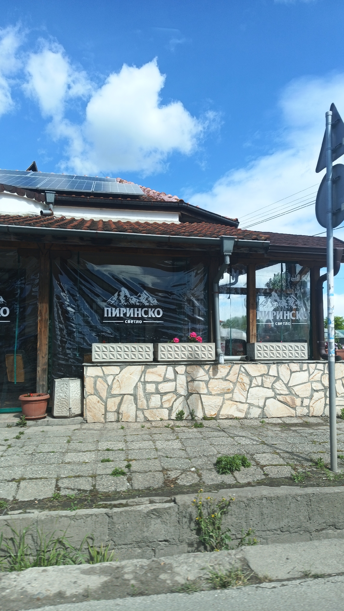 The image shows the exterior of a building in what appears to be Boliartsi, Bulgaria. The building has a visible sign reading "ПИРИНСКО" (PIRINSKO) with the word "светло" (svetlo) underneath, suggesting it is a restaurant or bar. The building features a tiled roof with solar panels, stone walls, and a covered patio area with transparent plastic sheeting bearing the PIRINSKO logo. The patio has concrete planters with pink flowers. The sidewalk in front of the building is paved with grey stones and shows some weeds growing in the cracks. The street is visible in the foreground, with a concrete curb. In the upper right corner, there is a glimpse of a road sign, suggesting it is located near a road intersection. The sky is blue with scattered white clouds.