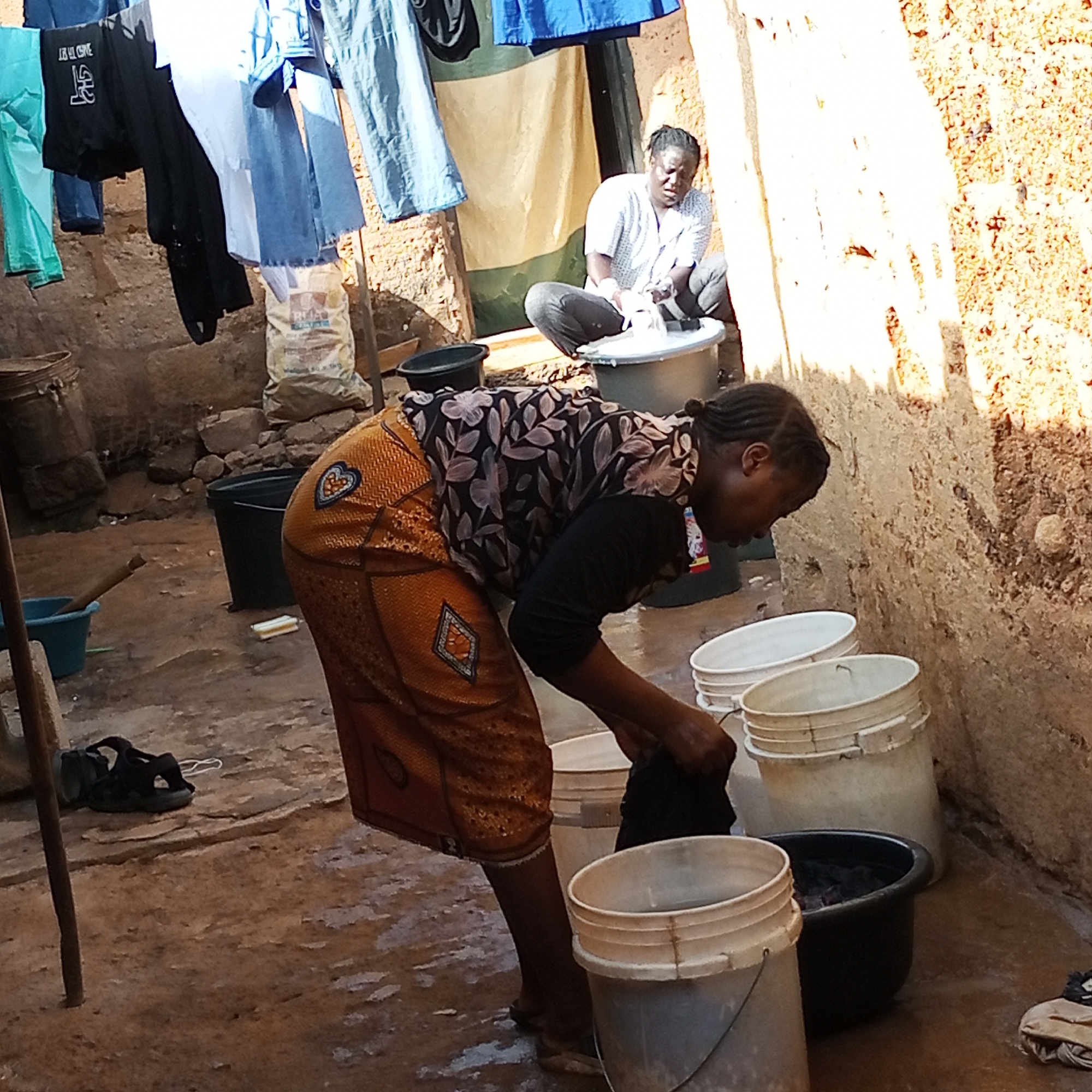 In an outdoor domestic setting in Jos, Nigeria, two women are engaged in laundry activities. The first woman, positioned in the foreground, leans over a collection of buckets, immersed in washing a dark piece of clothing. She wears a floral patterned top, a long orange patterned skirt, and sandals. The second woman, seated in the background, is washing clothes in a basin atop a bucket. Laundry hangs on a line behind her. The ground is wet and cluttered with buckets, footwear, and other objects. The surrounding walls appear to be made of mud or brick.