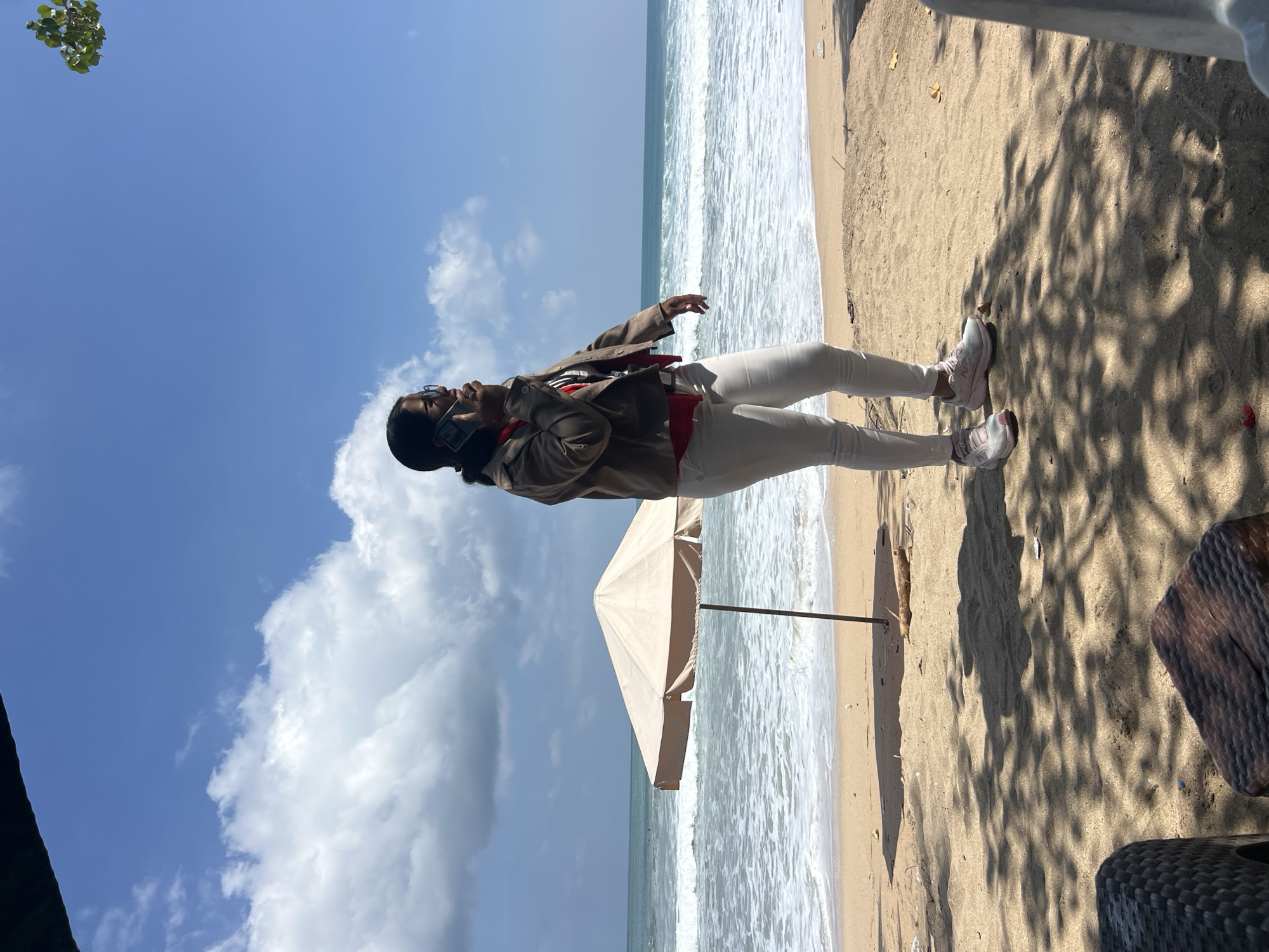 A full-body shot shows a woman in profile on a sandy beach, standing near the ocean and talking on her cell phone. She is wearing a brown jacket over a red shirt, white pants, and pink and white sneakers. She has dark hair pulled back and wears glasses. The beach has a beige umbrella, ocean waves, and a clear blue sky with some white clouds. In the foreground, there are two wicker chairs or ottomans partially visible in the shade of trees. The scene is set in Kuta, Indonesia.