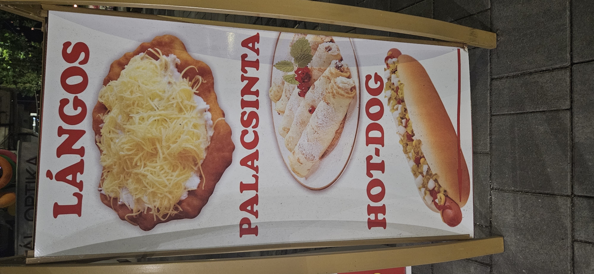 The image shows an A-frame sign advertising food items in Hévíz, Hungary. The sign has three sections, each featuring an image and text. The top section shows a Lángos, a traditional Hungarian fried dough dish, topped with cheese and sour cream. The middle section displays Palacsinta, Hungarian crepes or pancakes, dusted with powdered sugar and garnished with berries. The bottom section features a Hot-Dog, a sausage in a bun with various toppings. The sign is positioned on a paved sidewalk, with greenery visible in the background. The overall scene suggests an outdoor food vendor or a restaurant's promotional display.
