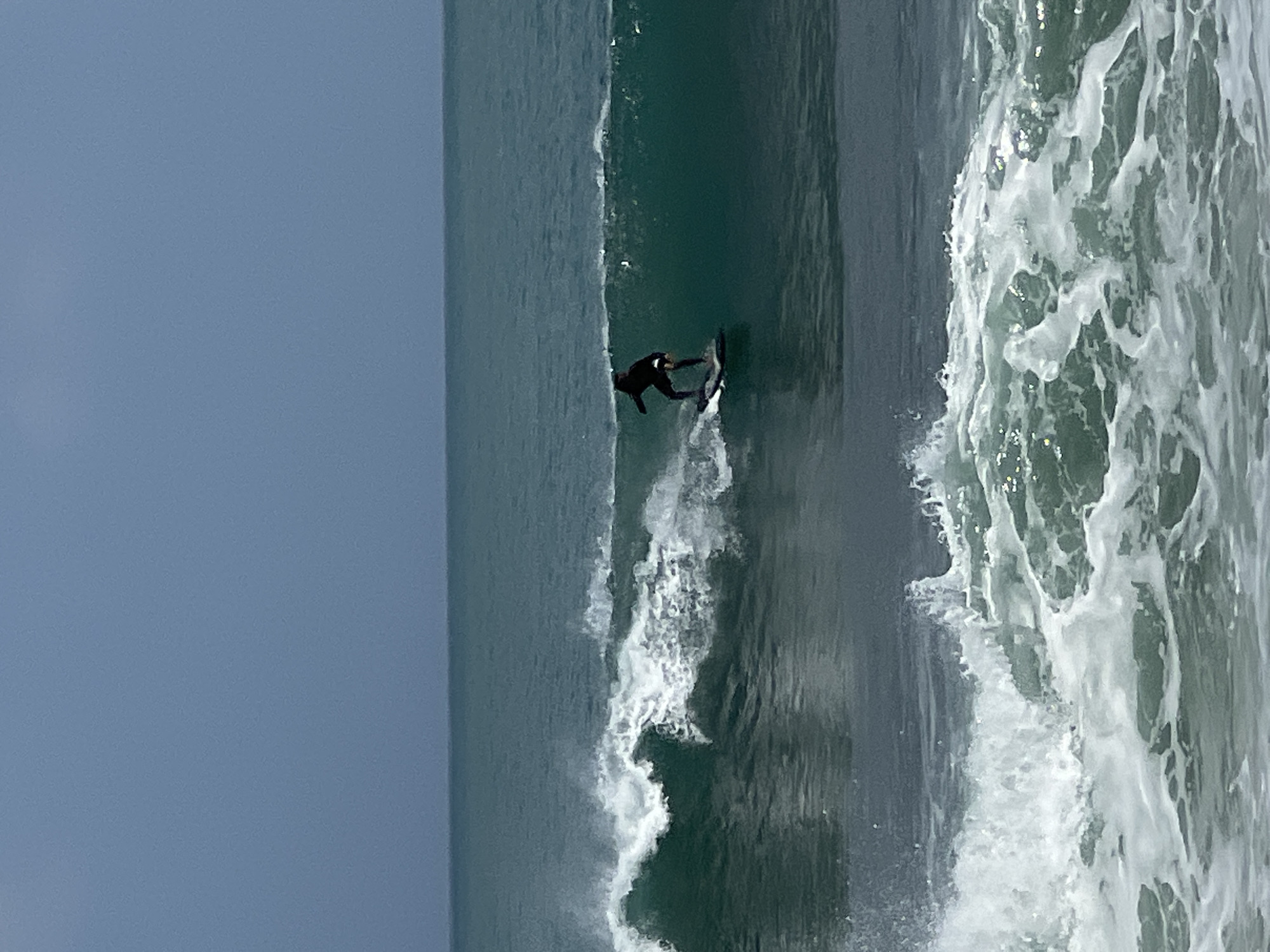 A lone surfer in a black wetsuit is riding a breaking wave. The surfer is positioned on the left side of the wave, with the wave curling over them. The water is a deep teal color, and the sky is a pale blue, suggesting an overcast or hazy day. In the foreground, another wave is breaking with white foam and spray. The scene appears to be a surf spot in Kuta, Indonesia, characterized by its consistent waves and popularity among surfers. The image captures a dynamic moment of surfing, with the surfer actively engaged with the ocean's power.