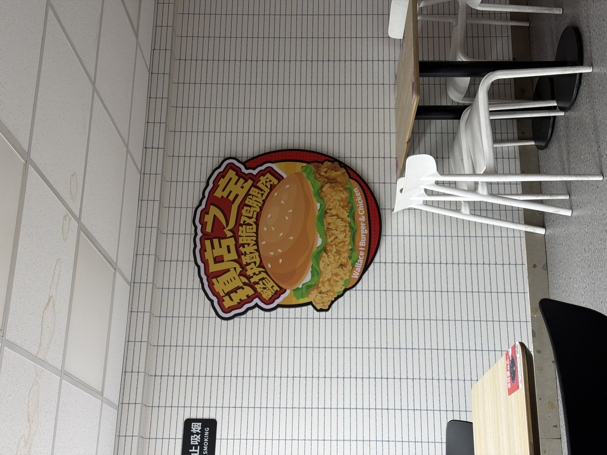 The image shows a wall in what appears to be a restaurant or fast-food establishment, as indicated by the "Wallace | Burger & Chicken" sign featuring a burger graphic in Chinese and English. Below the sign is a "No Smoking" sign in Chinese and English. The wall is constructed from white tiles with grey grout lines. To the right, tables and chairs are visible, suggesting a seating area. The presence of a table with a light wood-grain surface and modern chairs, some stacked, indicates a dining space. The overall scene is generic but informative. Specifics like the exact location within Jiangning District, China, or further details about people or interactions are not discernible from the image.