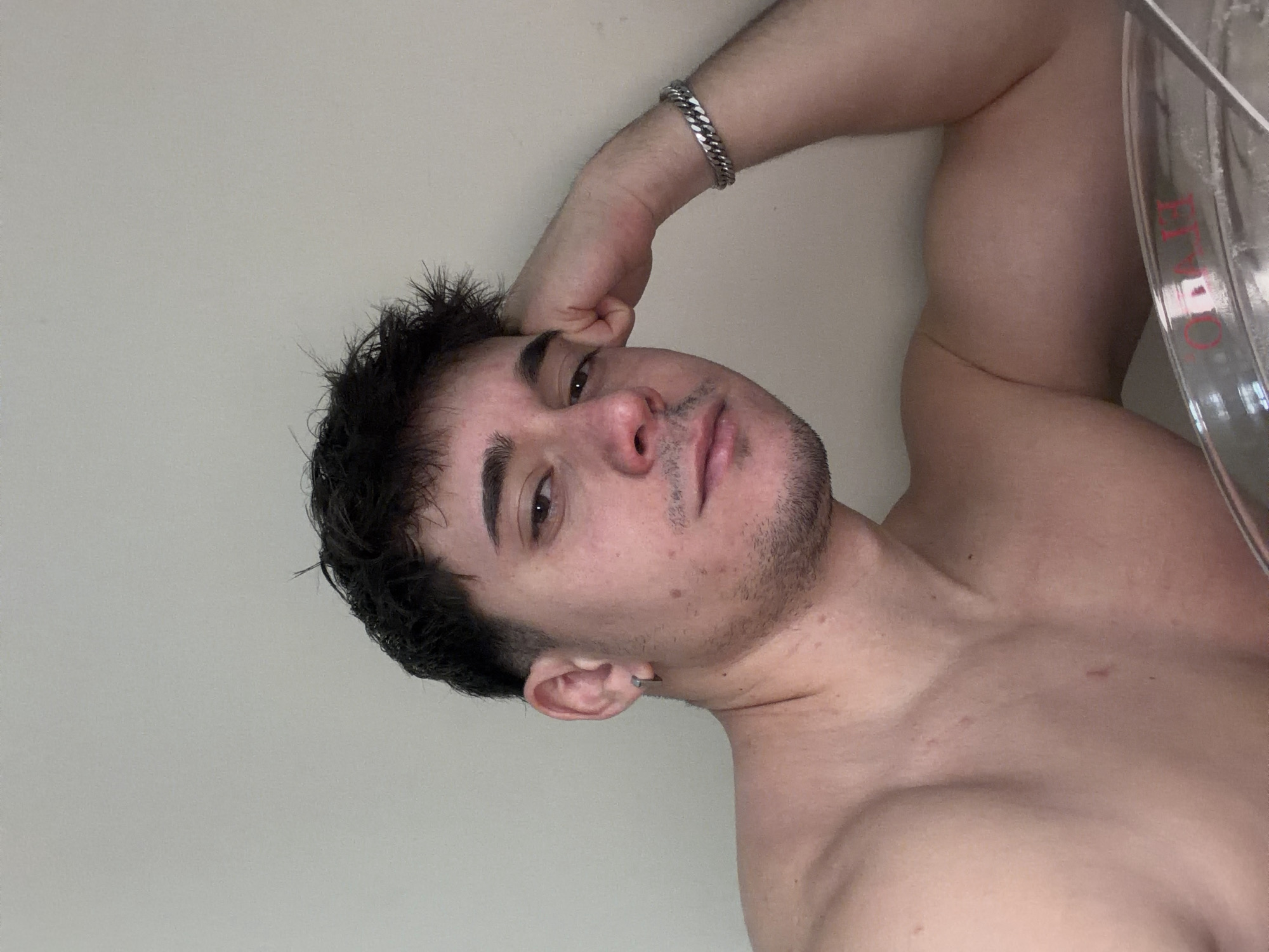 A shirtless man with dark hair and light skin poses in a room with a light-colored wall. He is lying on his side, with his head resting on his hand, which is adorned with a silver bracelet. He has stubble and a piercing in his ear. A transparent object with red lettering is partially visible on the right edge of the image. The specific location in Sofia, Bulgaria cannot be determined from the image.