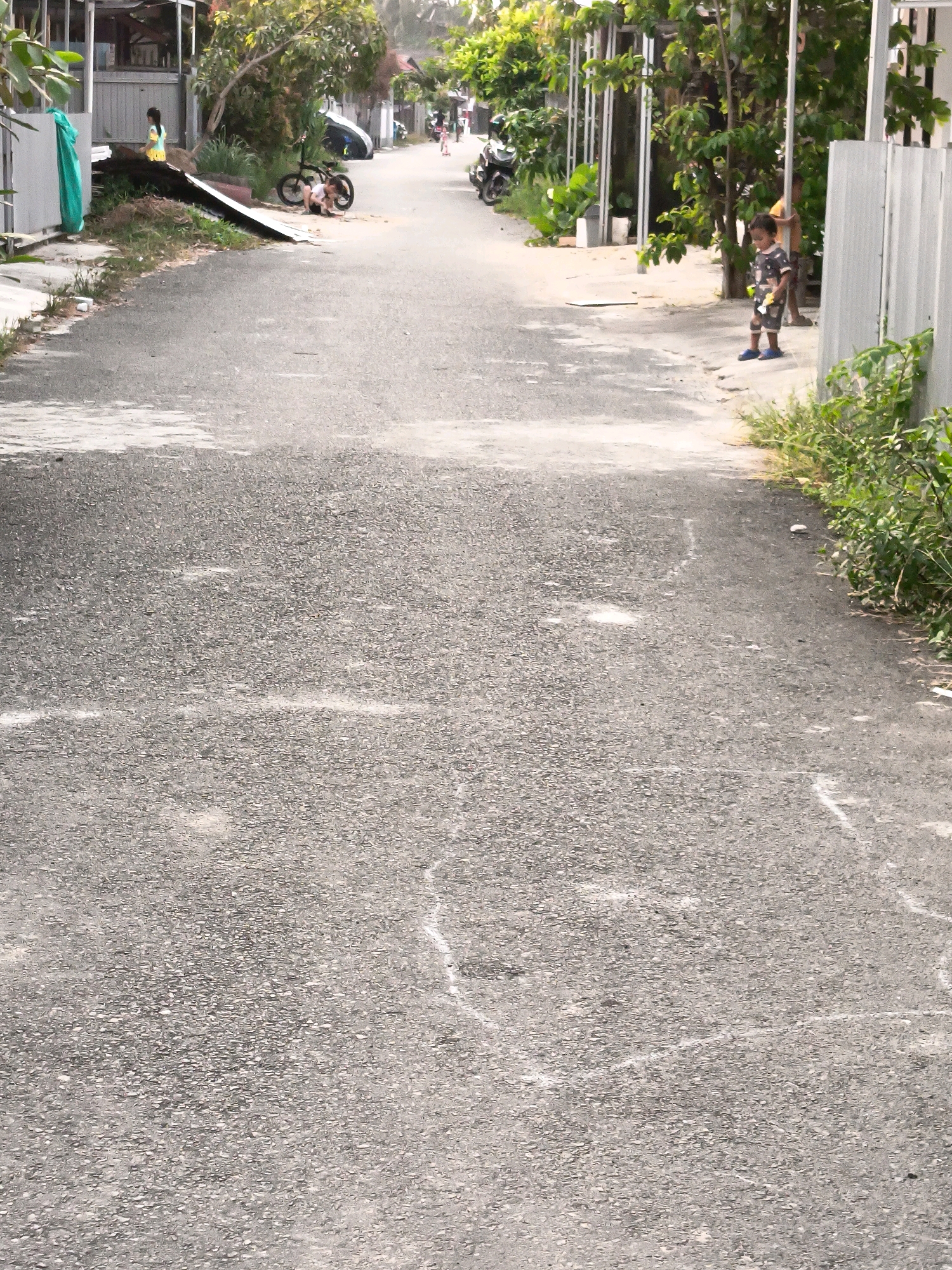 The image depicts a residential street in Banjarmasin, Indonesia. The street is paved with asphalt and shows signs of wear, with several white chalk lines visible on the surface, possibly indicating a game like hopscotch. On the left side of the street, a girl wearing a yellow and blue outfit stands near a pile of debris and a green tarp. Further down the street, a young boy in a camouflage t-shirt and blue sandals stands on the sidewalk. To the left of the boy, a motorcycle is parked. Trees and vegetation line the sides of the street, with houses and fences visible in the background. The overall scene suggests a casual, everyday environment in a residential area.