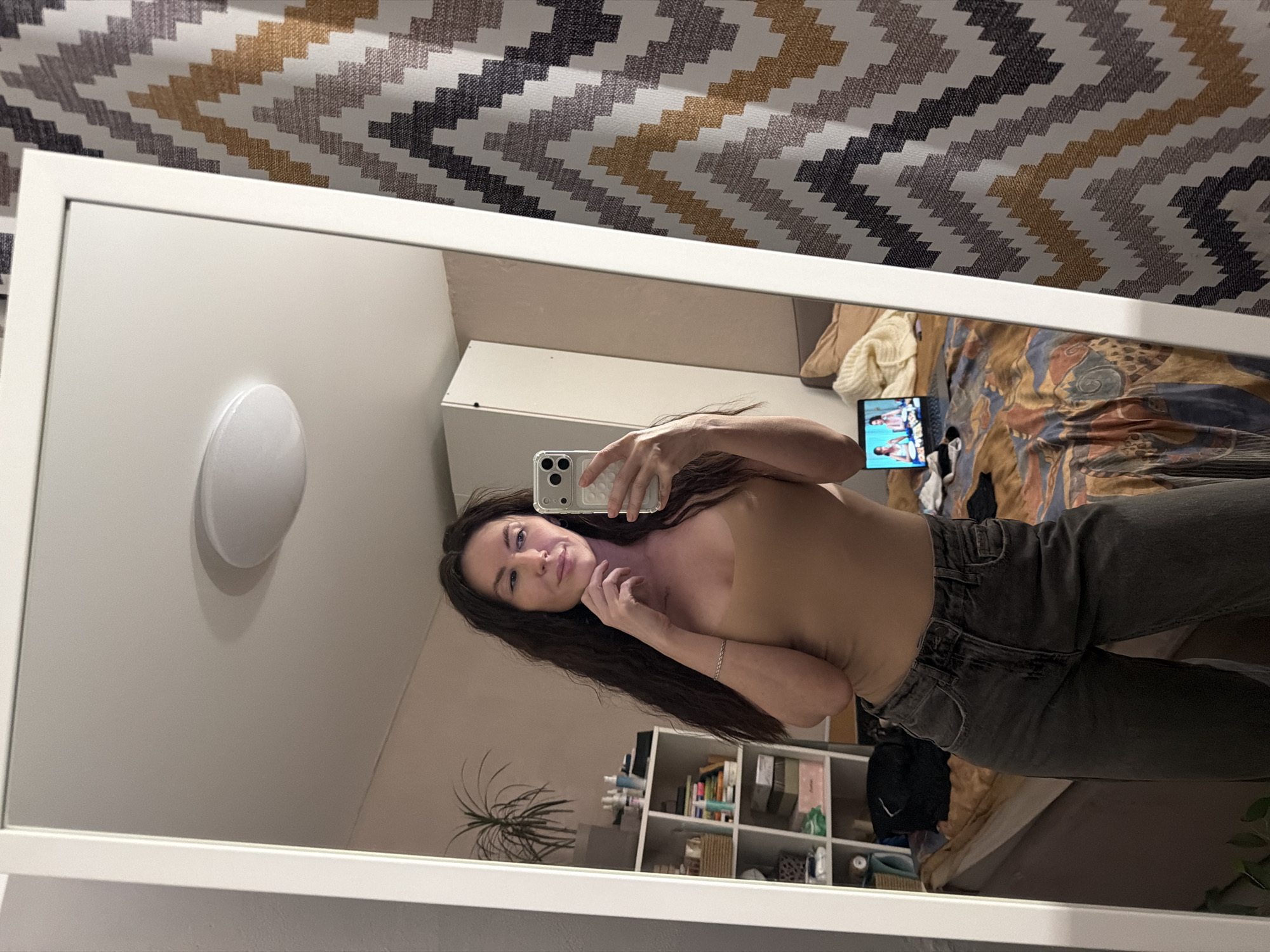 This image depicts a single adult female taking a mirror selfie in a residential interior within the city of Perm, Russia. The female subject has long, dark, wavy hair and is wearing a light tan sleeveless top and dark grey jeans. She holds a light-colored smartphone, visible with its camera array, in her right hand, positioned to capture her reflection. Her left hand is raised, touching her chin/jawline. A subtle silver chain bracelet is visible on her left wrist.
The mirror's reflection reveals a room with light-colored walls. To the left, a tall potted plant with green leaves stands next to a white open shelving unit stocked with various personal items including bottles, books, boxes, and woven baskets. Further right, a portion of a white wardrobe or cabinet is visible. A bed occupies the lower right portion of the reflected scene, covered by a patterned duvet featuring brown, orange, blue, white, and dark geometric designs. An open laptop rests on the bed, displaying two female figures on its screen. A beige throw blanket is draped over the bed. The ceiling features a circular, flush-mounted light fixture. Outside the mirror's frame, on the right side of the image, a patterned curtain with a zigzag or chevron design in shades of brown, beige, white, and dark grey is visible.