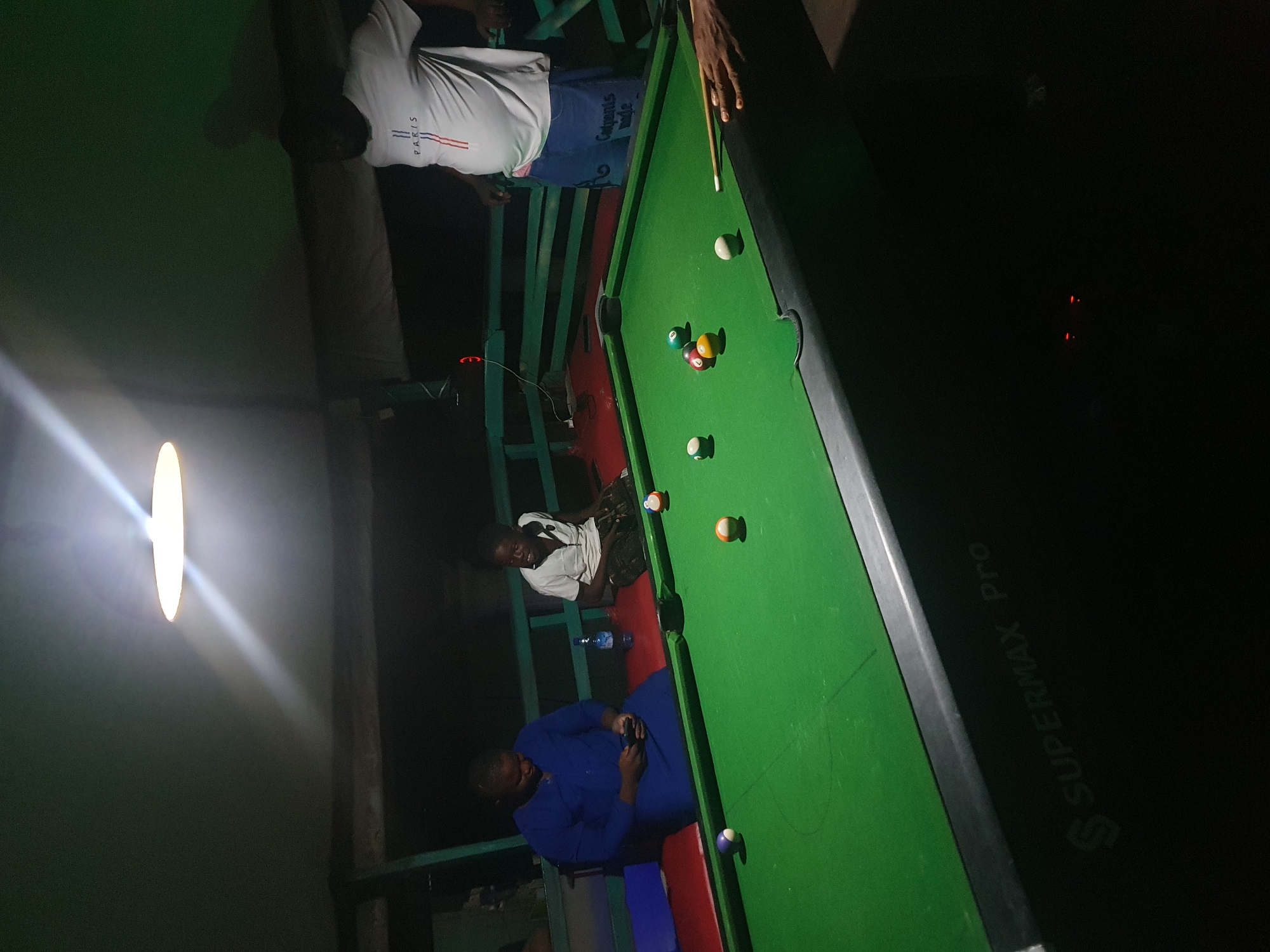 The image depicts a group of individuals engaged in a game of pool at an outdoor establishment in Lokoja, Nigeria. A pool table with several colored balls is centrally positioned. In the foreground, a person's hand is visible holding a pool cue, poised to strike a ball. To the left, two individuals are seated on a bench. One, wearing a blue garment, is looking at a mobile phone. Next to them, a young boy in a white shirt and patterned shorts is also seated, smiling and looking towards the table. To the right, another person, wearing a white t-shirt with "PARIS" printed on it and blue shorts, stands by the pool table. The environment appears to be a covered outdoor area, possibly a bar or recreational space, with dim lighting from an overhead lamp casting shadows. The presence of the "SUPERMAX Pro" branding on the side of the pool table suggests a specific type of equipment.