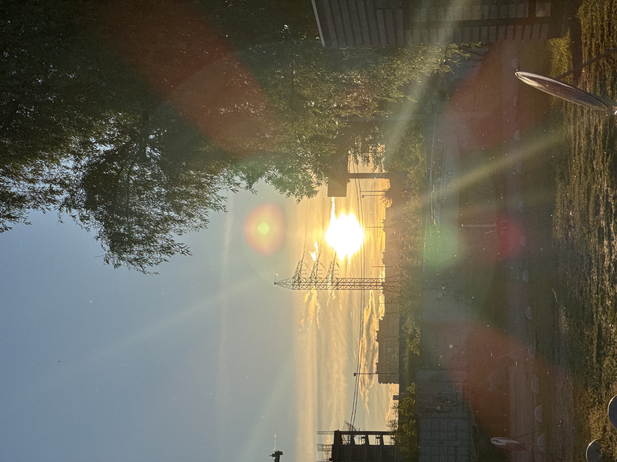 The image shows a sun setting over a city. In the center of the frame, a power transmission tower is silhouetted against the sun. Below the tower, there are buildings, some of which appear to be under construction. To the left and right of the tower, trees partially obscure the view. The sky is a gradient of colors, with the sun casting a bright glow. The foreground is somewhat blurred, with a hint of grass on the right side. There are visible lens flares and dust particles in the air.
