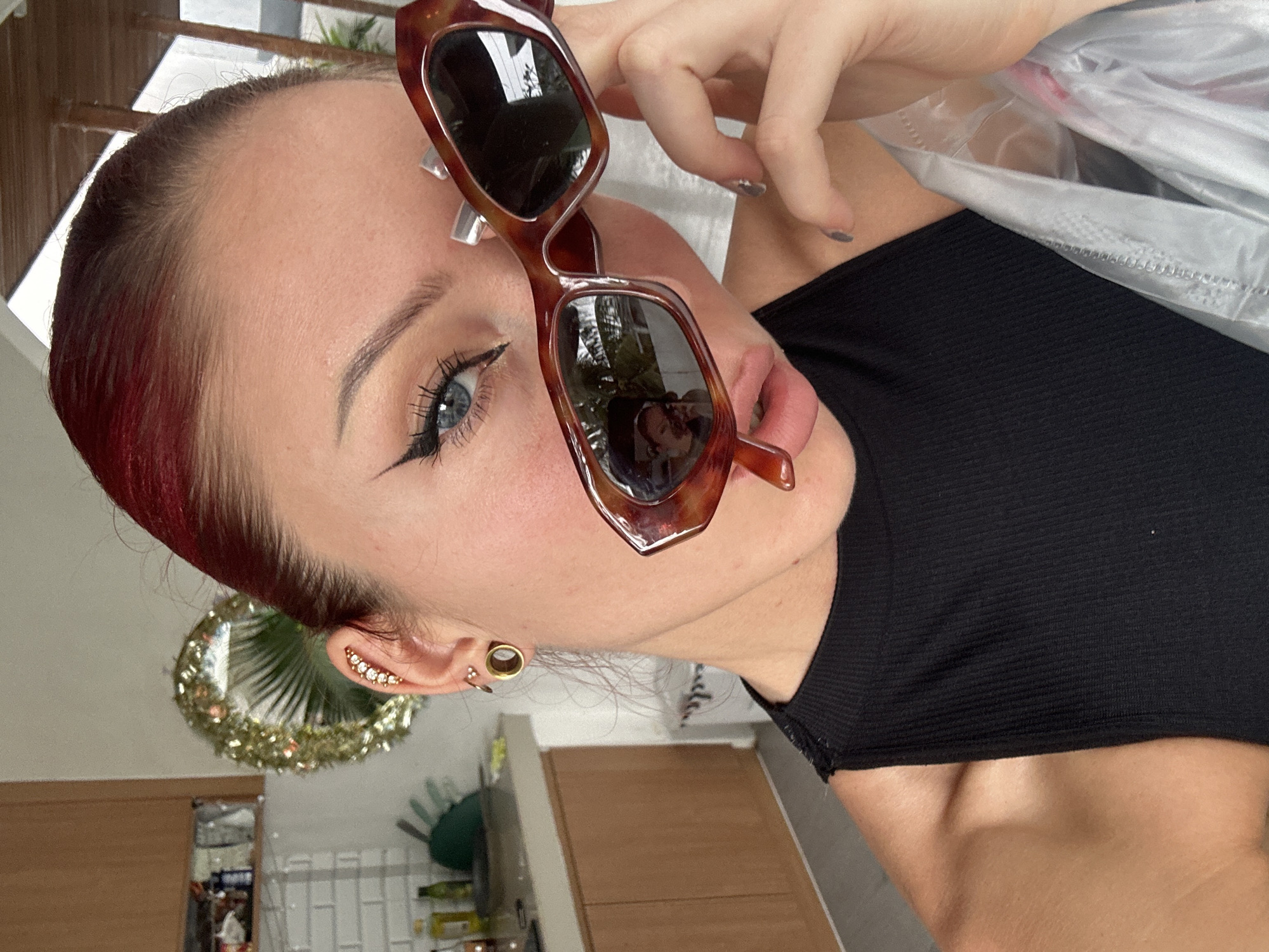 A close-up, vertically oriented photograph captures a person, likely a woman, posing in an indoor setting in North Kuta, Indonesia. The subject is holding a pair of brown tortoise-shell sunglasses above her eyes with her left hand, which has grey-painted fingernails. She has reddish-brown hair, styled back, and striking blue eyes enhanced with bold black eyeliner and long eyelashes. Her eyebrows are well-defined, and her lips are slightly parted.
She is wearing a black ribbed tank top or crop top. Her visible ear features multiple gold-toned piercings: an ear cuff with a line of gems along the outer helix, a prominent tunnel or gauge in her lobe, and another smaller piercing further up.
The background suggests a home environment, possibly a kitchen or living area, with visible wooden cabinetry, a white tiled backsplash, and various bottles and a green bowl. A large, circular, golden and sparkly decoration, resembling a wreath or tinsel, hangs in the background. Natural light illuminates the scene, indicating it is daytime. A clear plastic bag is partially visible in the upper right portion of the frame. The subject's expression is neutral to slightly pensive or alluring as she looks towards the camera. There is no visible text in the image.