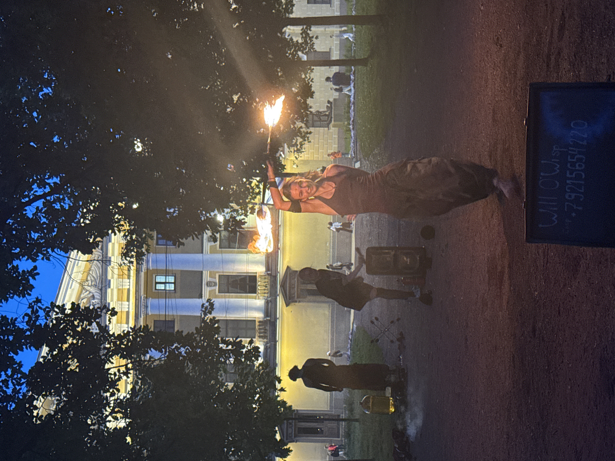 A young woman performs fire dancing in front of the Stroganov Palace on Nevsky Prospekt in Saint Petersburg, Russia. The palace, lit by exterior lights, is visible in the background, its yellow facade and classical columns contrasting with the dark foliage of trees. The woman, wearing a dark tank top and flowing skirt, twirls a fire prop, emitting flames and sparks. To her left, a dark, silhouetted figure stands near a large yellow container. In the foreground, a dark sign with white lettering, likely a name and phone number, is propped up. The ground is a mixture of gravel and dirt, and the sky is a deep blue, indicating dusk. Other figures are visible in the background, some walking and others seated, suggesting a public gathering.