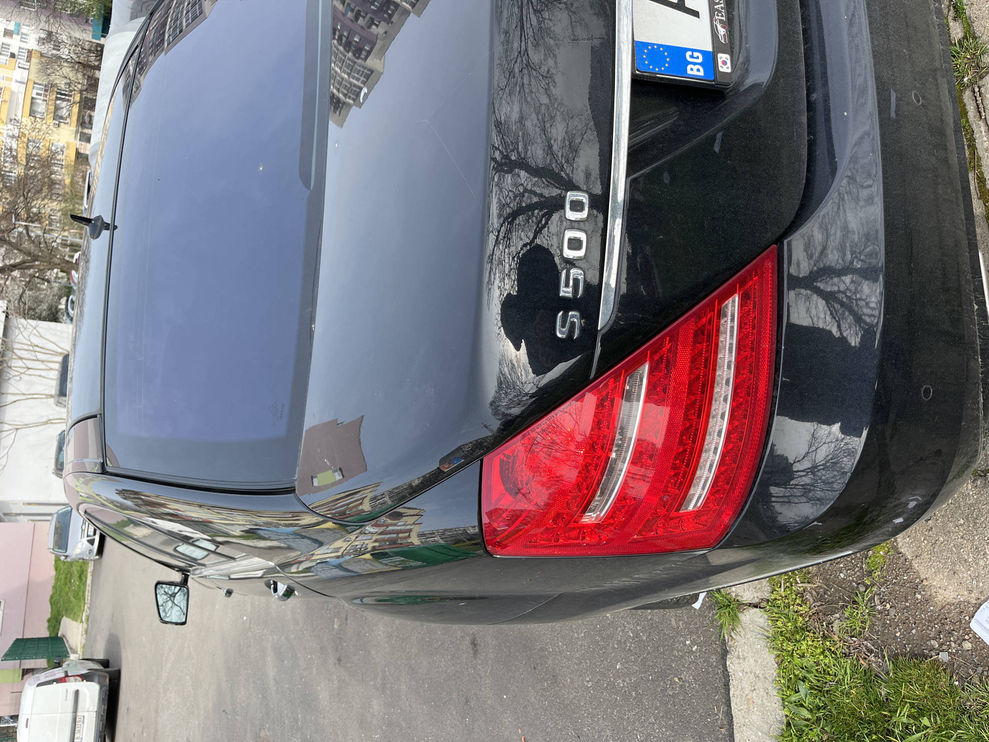 The image shows the rear of a black Mercedes-Benz S500 parked on a street in what appears to be a residential area. The car has a Bulgarian license plate (BG). The location appears to be in an urban setting based on the buildings reflected in the car's surface. There's a taillight visible on the right side of the frame, and the "S500" badge is clearly visible on the trunk. The vehicle is parked next to a curb with some grass and debris. There is no evidence indicating that the image was captured in Burgas, Bulgaria.