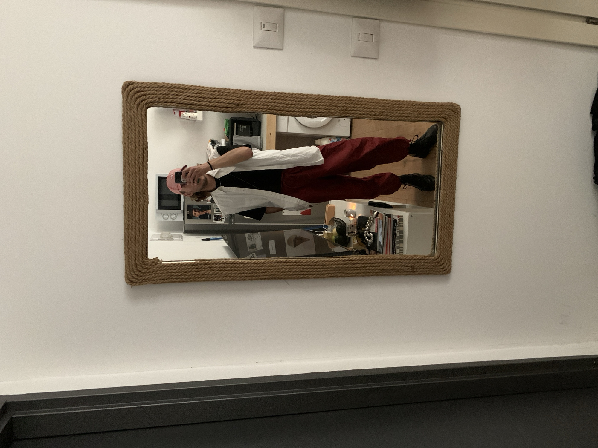 The image shows a mirror with a rope frame hanging on a white wall. Reflected in the mirror is a person wearing a pink hat, a black t-shirt, a white shirt, and red pants. They are holding a phone and taking a picture of themselves. The reflection also shows a room with a washing machine, microwave, a shelf with books, and other items. The scene appears to be indoors, possibly in an apartment. No specific location in Tel-Aviv, Israel is discernible from the image.