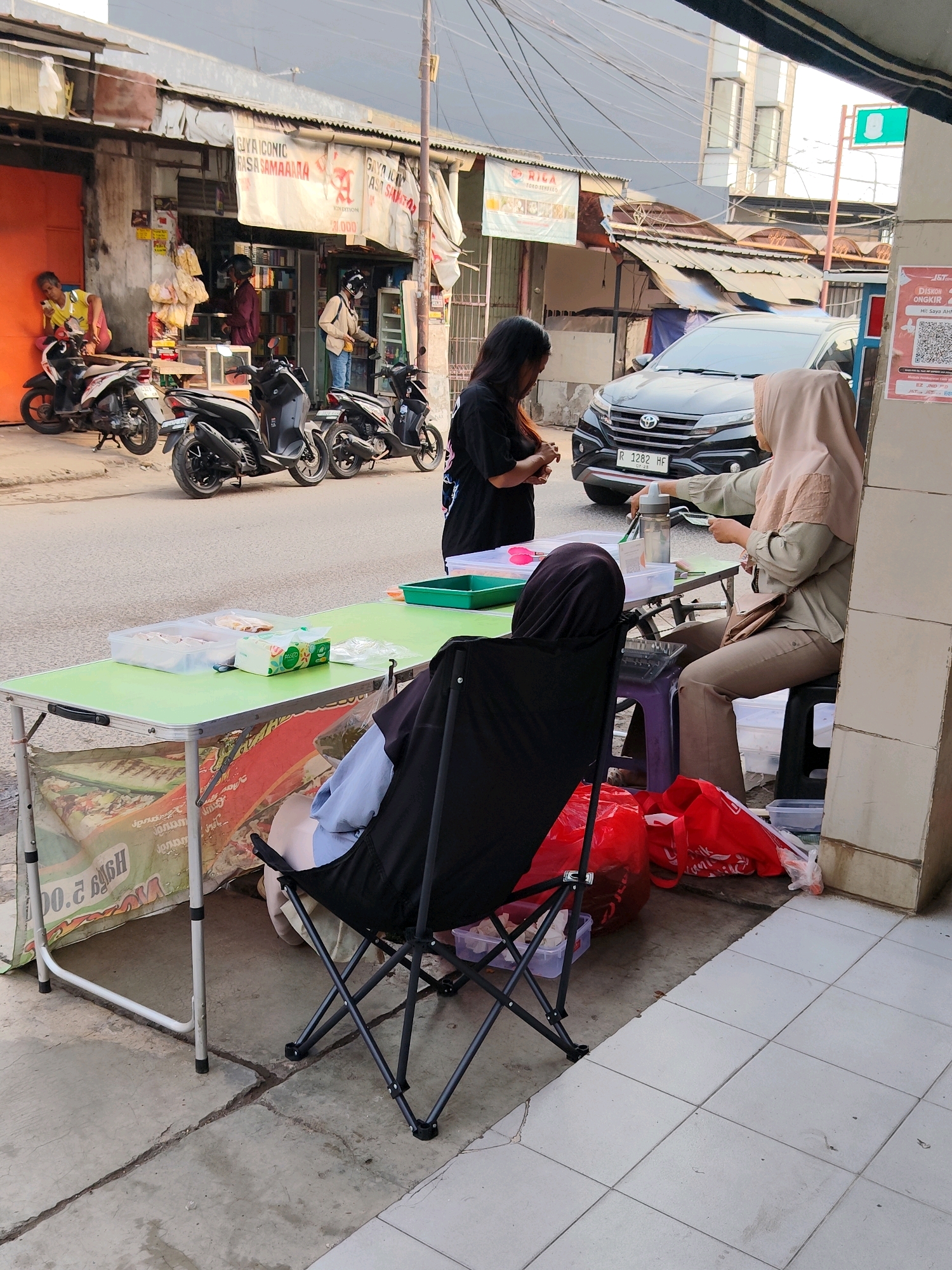 The image depicts a street scene in Bekasi, Indonesia, likely a commercial area with a row of shops lining the street. In the foreground, a green folding table is set up on the sidewalk, displaying a box of tissues and a plastic container with what appears to be food items. A person wearing a black headscarf is seated in a folding chair behind the table, facing away from the camera. To the right of the table, another person, a woman wearing a light brown hijab and a green top, is seated on a stool, interacting with items on the table. A black car with the license plate "R 1282 HF" is parked on the street behind them. Several motorcycles are parked along the curb in front of the shops. The shops themselves have awnings and display various goods, suggesting a market or retail environment. The architecture is functional, with corrugated metal roofs and simple facades. The overall scene suggests a bustling, everyday street life in an Indonesian city.