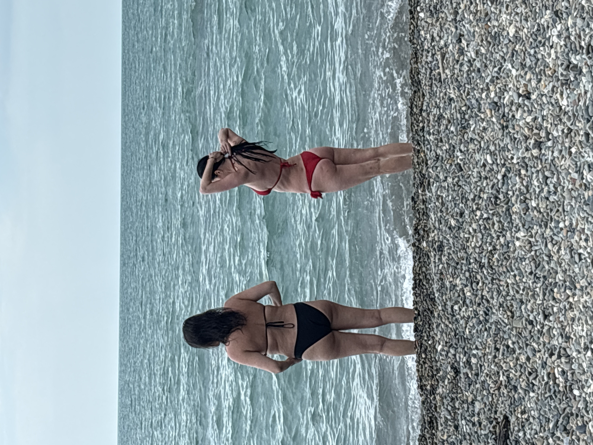 The image shows two women in bikinis at a pebble beach in Sochi, Russia. The woman in the foreground is wearing a black bikini and is facing away from the camera with her hands on her hips. The woman in the background is wearing a red bikini and is also facing away from the camera. She is touching her hair. Both women are standing in the shallow water of the sea. The sea is a light blue-green color with gentle waves. The beach is covered in small, grey pebbles. The sky is a pale blue, suggesting it is a clear day. The image appears to have been taken during daylight hours, likely in the afternoon given the bright light. There is no visible text in the image.