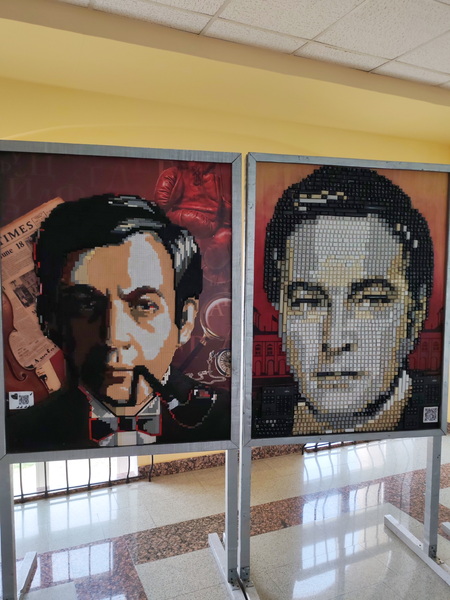 Two framed artworks, each depicting a portrait, stand on a white metal frame structure. The portrait on the left depicts a man in a dark suit smoking a pipe, against a reddish-brown background with a newspaper, boxing gloves, and a pocket watch. The portrait on the right is composed of keyboard keys and depicts a man's face with a building visible in the background. Both artworks include QR codes. The floor has a marble or tiled pattern. The walls are yellow and there is a tiled ceiling. The location is in the city of Agryz, Russia.