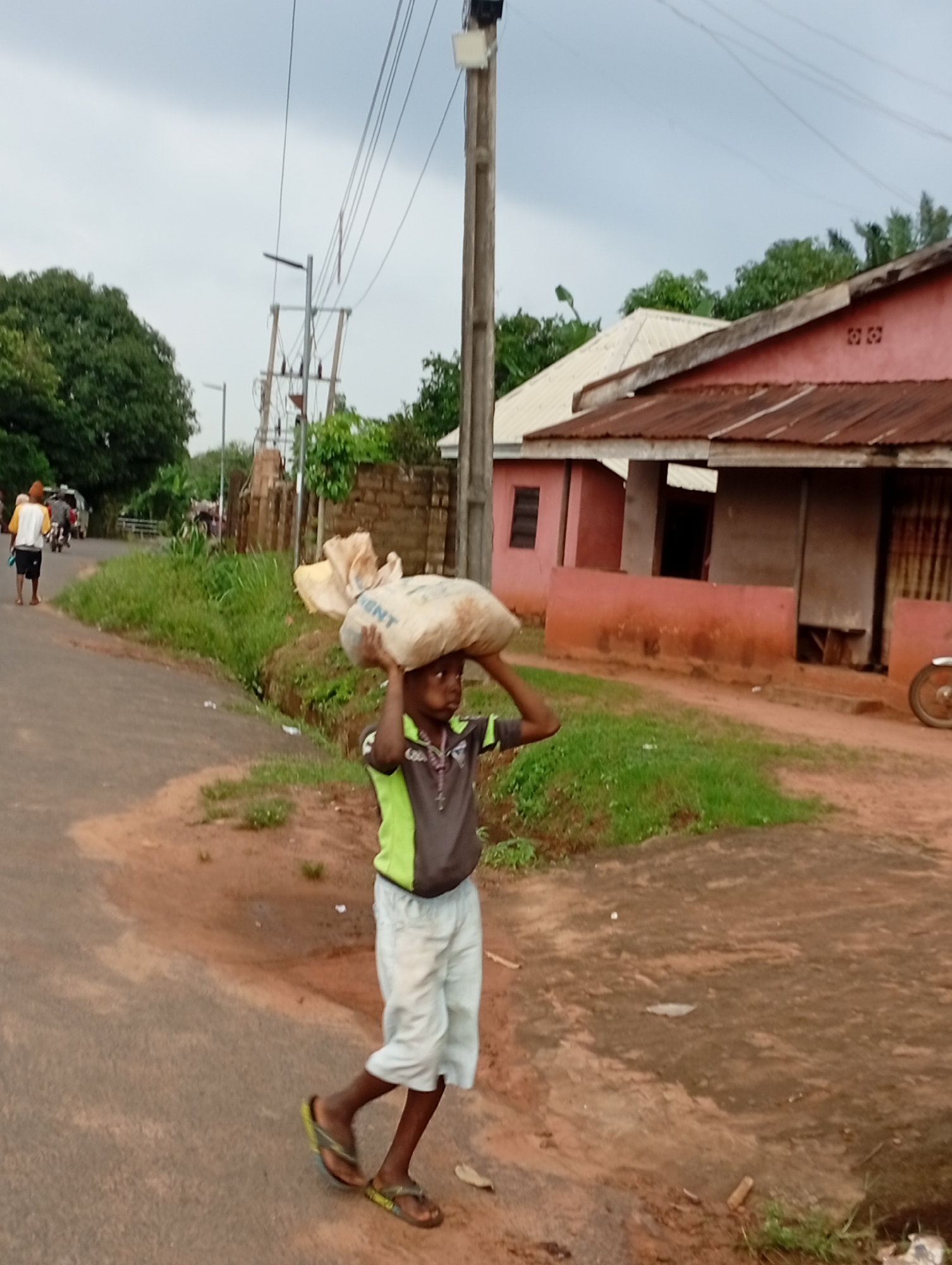 A young boy walks along a paved road in Nsugpe, Nigeria, carrying a large sack of goods on his head. He is wearing a grey and lime green t-shirt and light-colored shorts. His feet are clad in flip-flops. The road is flanked by sparse vegetation and a red-colored building with a corrugated metal roof. In the background, there are more buildings, trees, and utility poles with overhead wires. Another person, wearing a yellow top and dark shorts, is walking away from the camera on the same road, further in the distance. Vehicles are also visible on the road in the background. The sky is overcast.