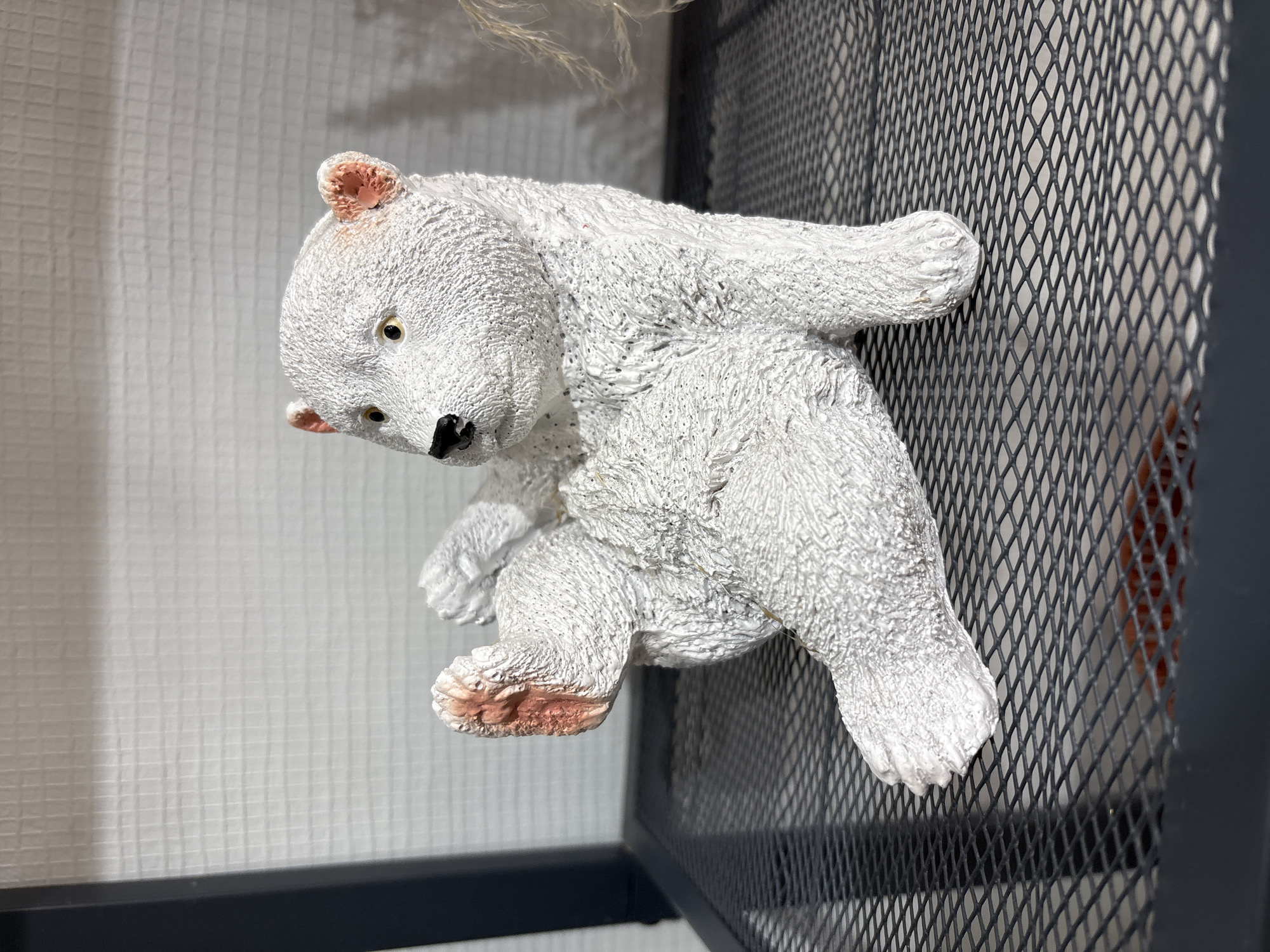 A white, textured polar bear figurine clings to the side of a black metal grid shelf. The bear has pinkish-tan accents on its ears and paws. The shelf is positioned against a textured, light grey wall. The location is not provided in the image, so the requested location information can not be included.