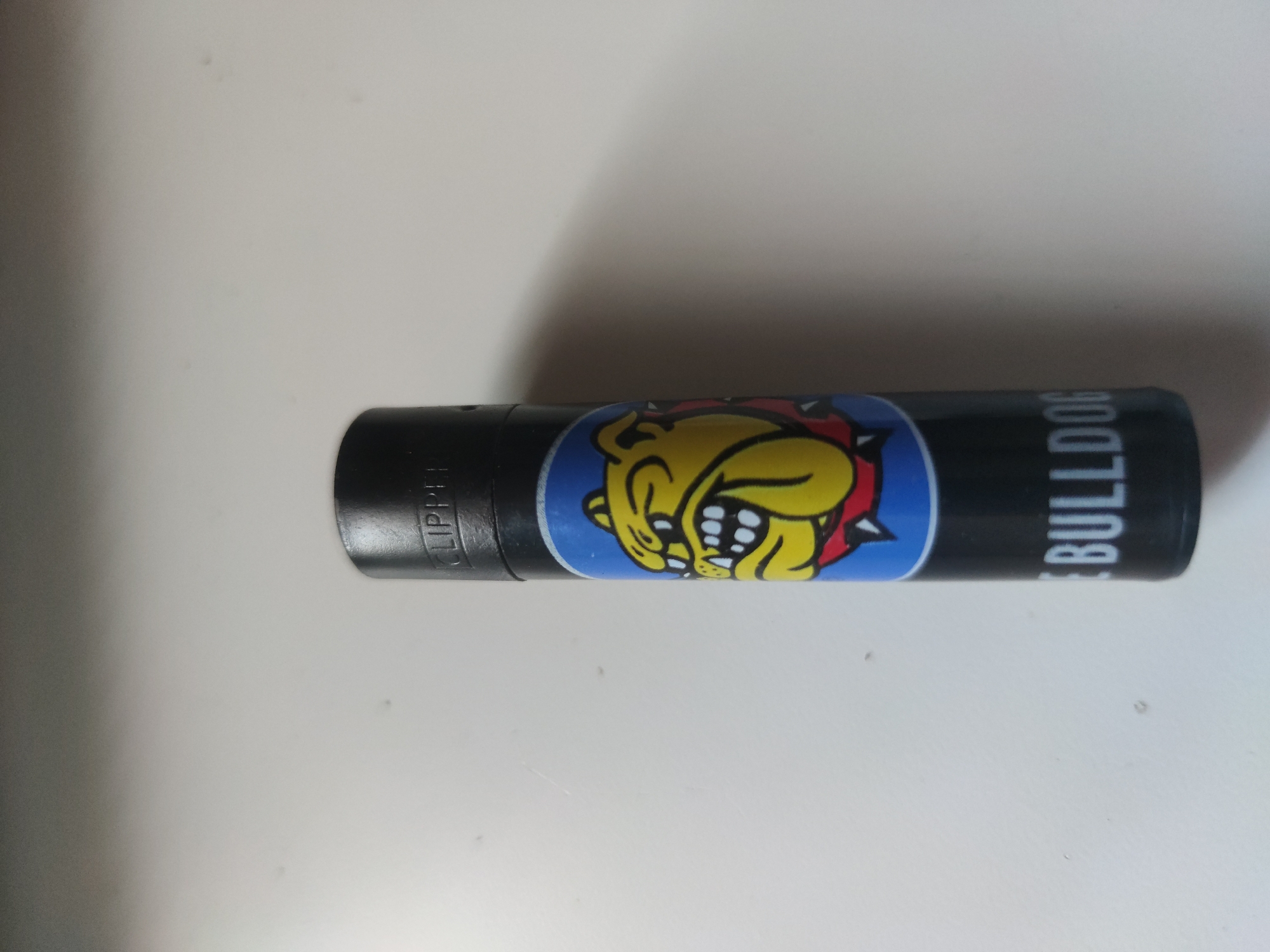A black Clipper lighter with a bulldog cartoon on a blue background is lying on a white surface. The word "BULLDOG" is printed on the lighter next to the bulldog cartoon. The location is unknown.
