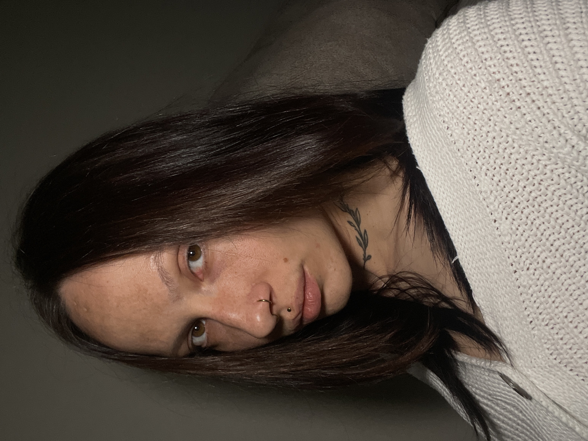 A brunette woman with a neck tattoo is lying down, wearing a white knit sweater. She has brown eyes, a nose ring, and a lip piercing. The background is a dimly lit gray wall. There is no evidence to suggest the location is Izhevsk, Russia.