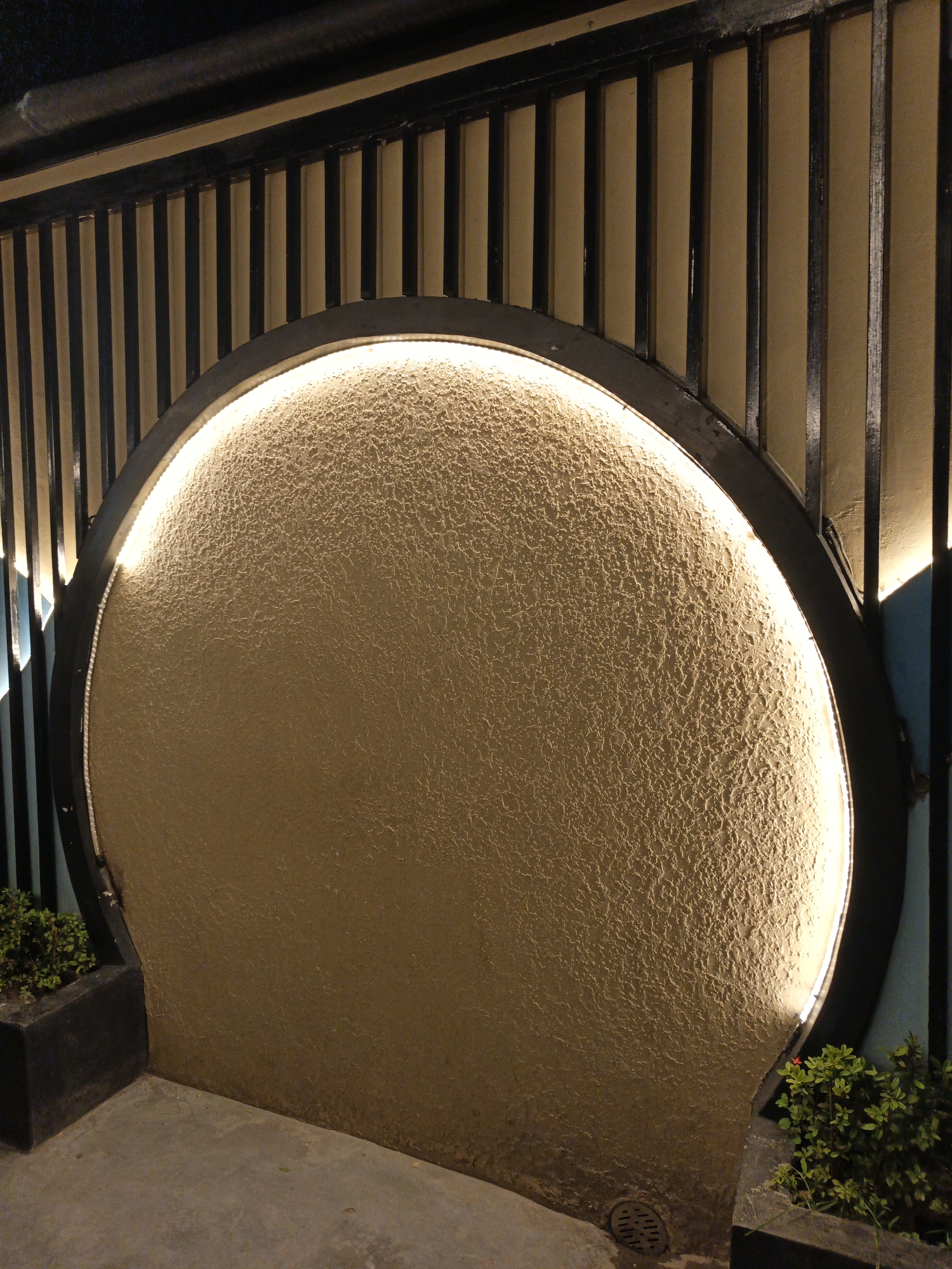 The image shows a circular opening with a textured, light-colored surface, illuminated from behind by a warm light source. The opening is framed by a dark, circular border. Behind the opening, a fence-like structure is visible, composed of vertical dark bars set against a light-colored background. Small, dark green plants in dark rectangular planters are positioned at the bottom left and right of the circular opening. The ground in front of the opening appears to be concrete or paving stones. The overall scene suggests an outdoor setting, possibly an architectural element in a garden or public space. No people are present in the image. The specific location in Dhaka, Bangladesh, cannot be determined from the visual information provided.