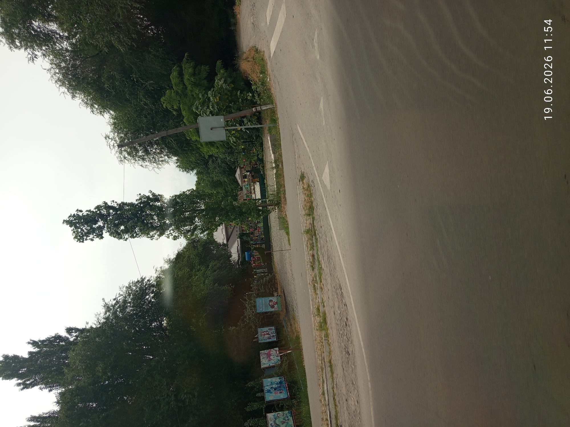 A dirt road curves to the right, meeting a paved road. Several signs, some with cartoon images and one in Russian reading "Ясли-сад Чебурашка," are posted along the dirt road, leading to a fenced area with playground equipment. Tall trees and foliage surround the area, and the sky is overcast. A wooden utility pole stands near the intersection. The date and time "19.06.2026 11:54" are visible in the lower right corner.