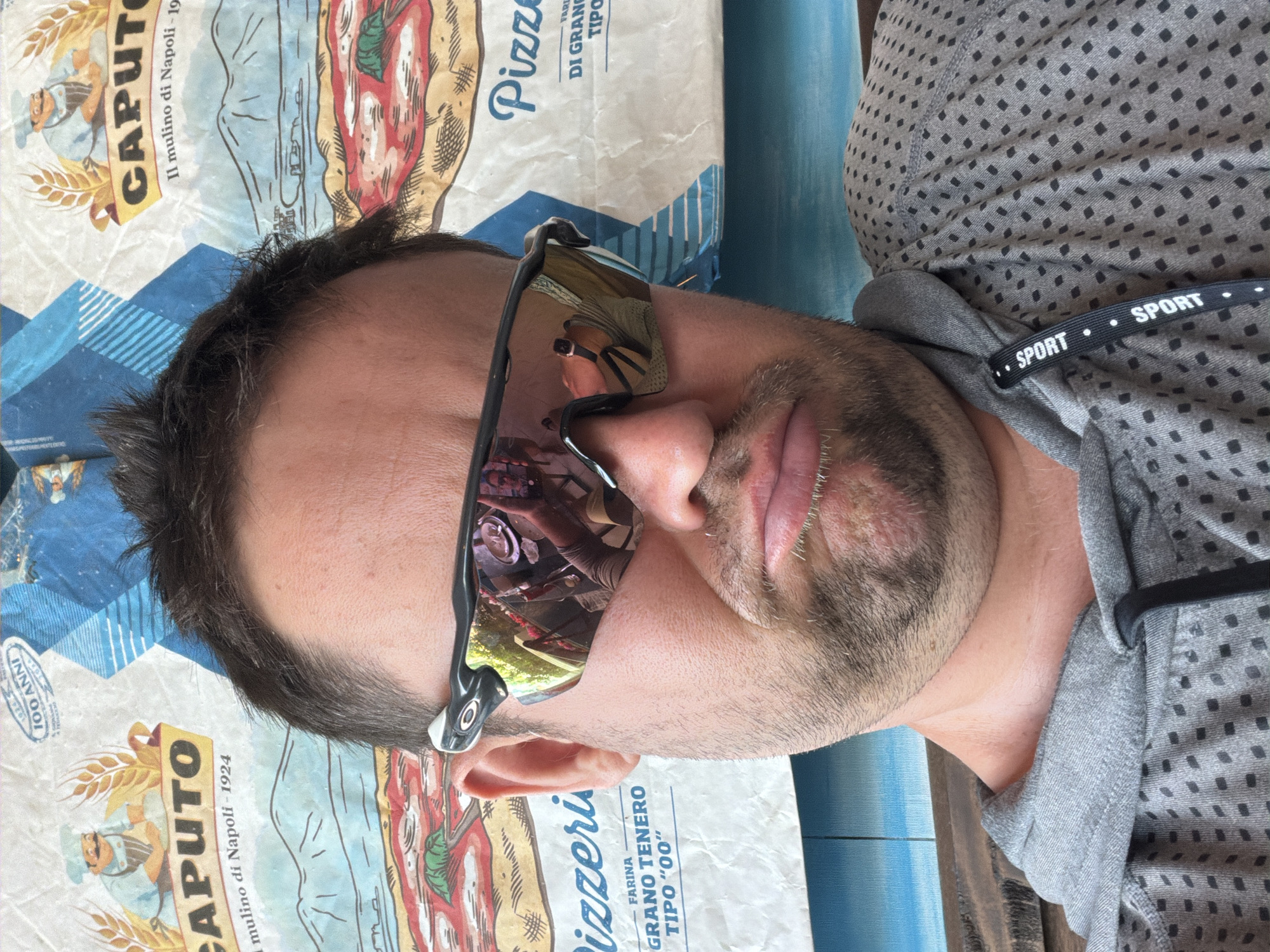 The image is a close-up selfie of a man wearing sunglasses and a grey hoodie. He is seated outdoors, as indicated by the background, which appears to be a wall decorated with a print of a pizza and text related to "Caputo" and "Pizzeria". The man has short, dark hair and a beard, and his mouth is pursed in a pout. The sunglasses reflect the surrounding environment, showing a glimpse of what appears to be tables and chairs, suggesting he is at an outdoor seating area of a restaurant. The text on the background print includes "CAPUTO", "Il mulino di Napoli - 1924", "FARINA GRANO TENERO TIPO '00'", and "Pizze". The hoodie has a repeating pattern of small dark squares on a grey background, and a black ribbon with the word "SPORT" printed in white is attached to the neckline. The lighting suggests it is daytime. There is no indication of the city of Baan Bo Phut, Thailand in the image.