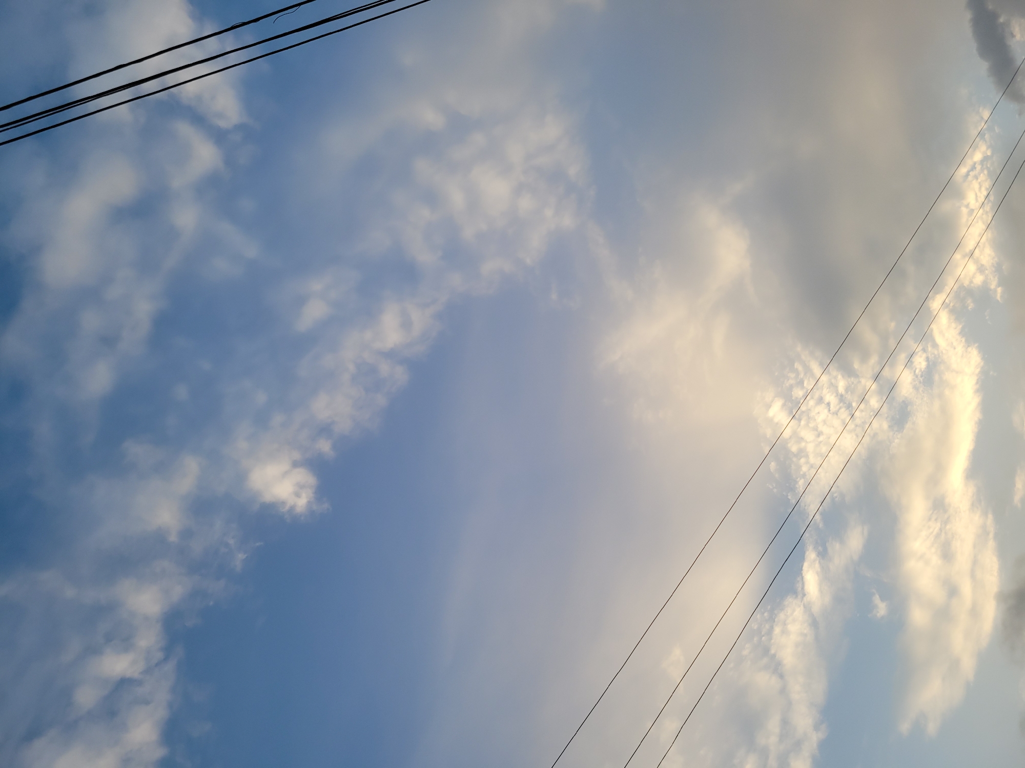 The image shows a sky with scattered clouds and three dark, thin power lines running diagonally from the top left to the bottom right. The sky is a pale blue, with some areas appearing whiter due to the clouds. The clouds themselves vary in density and color, with some appearing bright white and others a muted gray, especially towards the right side of the image, suggesting the presence of more substantial cloud cover or a different light angle. A warm, golden light is visible in the upper right quadrant, illuminating some of the clouds and creating a gradient effect in the sky. This suggests that the sun is either rising or setting, or is low on the horizon. There are no people or distinct objects visible in the scene, only the sky and the power lines, which are a common man-made element found in urban or semi-urban environments. The overall impression is one of a serene, perhaps transitional moment in the day, captured in an outdoor setting. The location context of Jos, Nigeria, is consistent with such a sky and the presence of power lines.