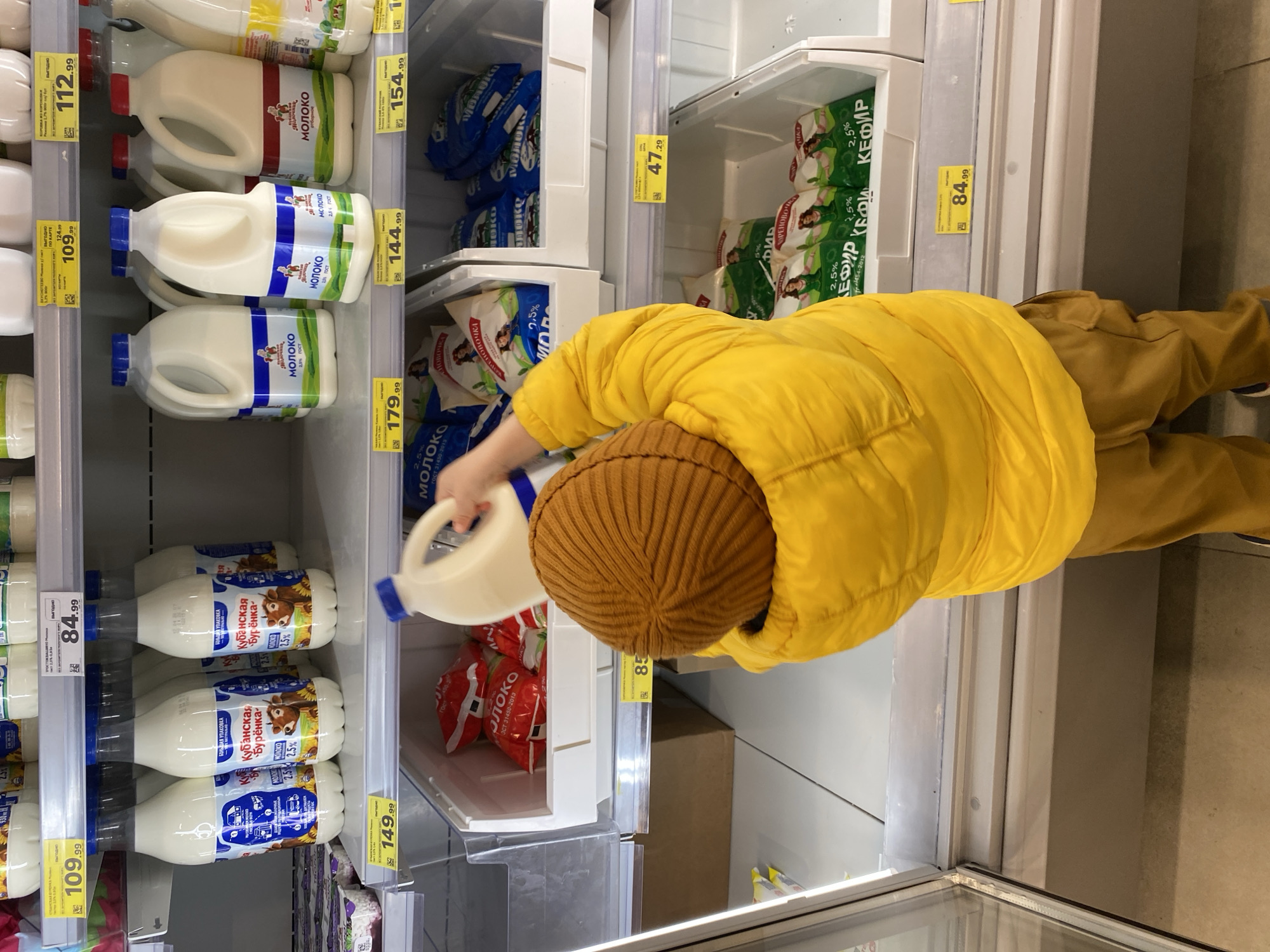 A young child wearing a brown knit hat and a bright yellow puffer jacket is reaching into a refrigerated dairy case in a grocery store. The child's right hand is grasping the handle of a large, white plastic jug of milk. The dairy case is stocked with milk and kefir products in bags and plastic jugs. The price tags on the shelves show the products are available for sale. The milk jugs have labels in Russian that translate to "Milk." The kefir packages are labeled with "Kefir." The prices are in rubles. This scene is most likely in a grocery store in Russia, possibly in the city of Sochi, given the Russian language labels on the products.