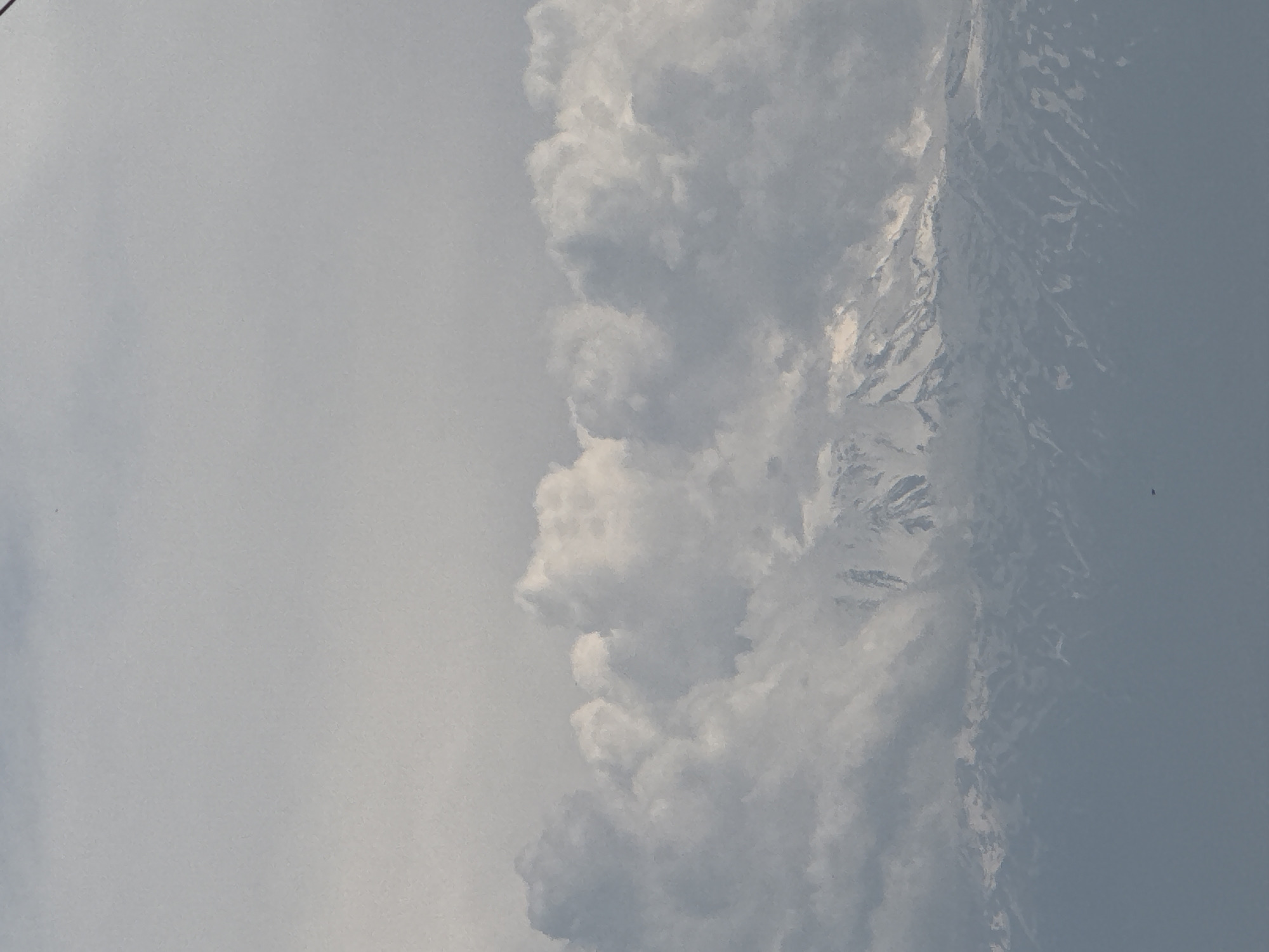 The image displays a vast natural landscape, appearing to be rotated 90 degrees clockwise from its intended orientation. When viewed correctly as a landscape, the scene features a expansive sky, a significant cloud formation, and a snow-capped mountain range.
The upper portion of the image shows a clear sky, transitioning from a light grey-blue at the top to a slightly darker tone as it approaches the cloud line. A large, prominent band of white and grey clouds stretches horizontally across the middle of the frame, thick and fluffy, partially obscuring the view below. Beneath these clouds, the rugged peaks of a mountain range are visible. These mountains are covered in snow, creating bright white patches that stand out against the darker rock and the atmospheric haze. The intricate textures of the snow and rock formations are discernible despite the distance and partial cloud cover.
The scene depicts a natural, untouched environment. No human figures, man-made objects, or visible text are present. The lighting suggests it is daytime, with the clouds contributing to a soft, diffused light over the landscape. The presence of snow indicates either a high altitude, a cold climate, or a winter season. The overall impression is one of majestic natural beauty and serenity.
