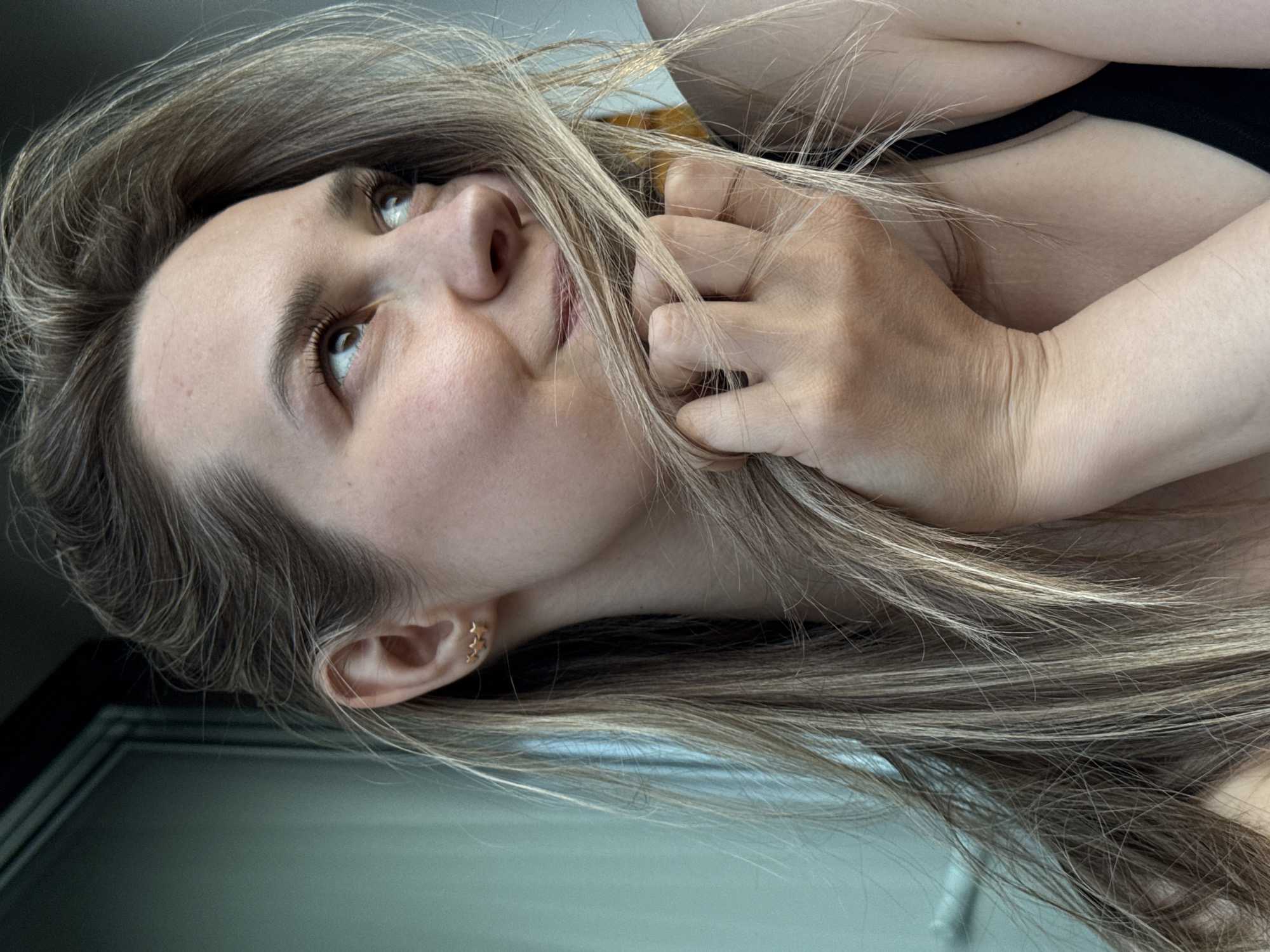A woman with light brown and blonde hair is looking upwards with a slight smile. Her hand is near her face, fingers partially intertwined with her hair. She is wearing a black top. A door frame is visible in the lower left corner. The location is not discernible.