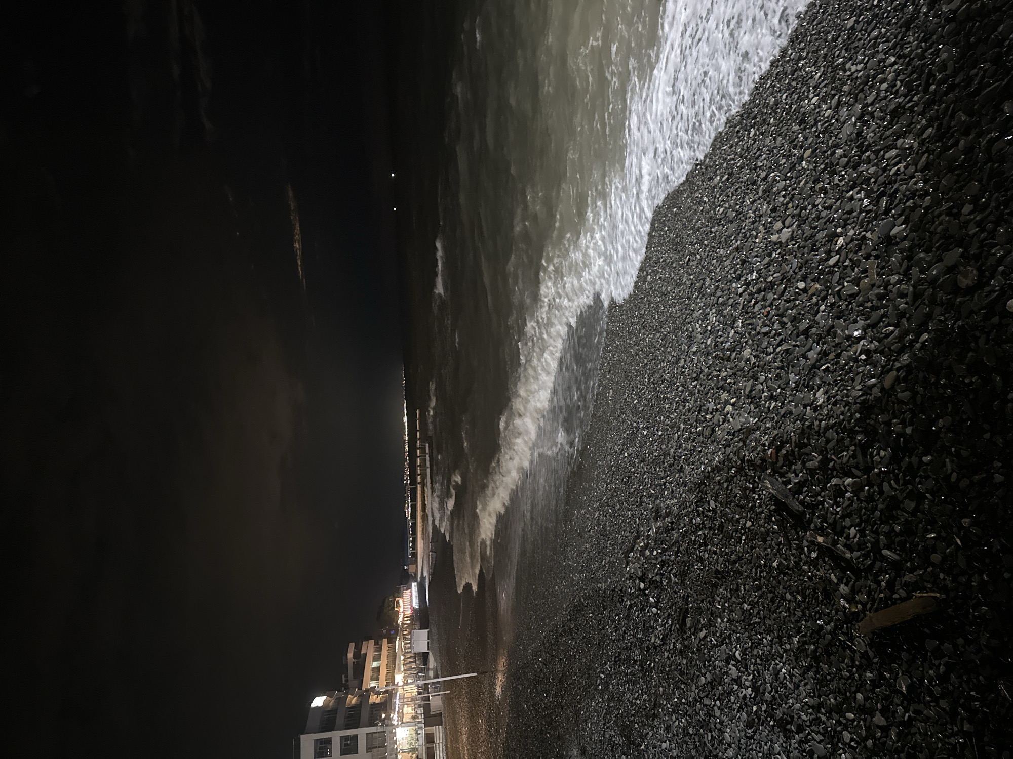 The image shows a night scene at a beach in Sochi, Russia. The shoreline is composed of dark pebbles. Waves are crashing onto the beach, illuminated by artificial lights coming from buildings in the background. The sky is dark, and some buildings are visible on the left side of the frame. Overall the light is dim.