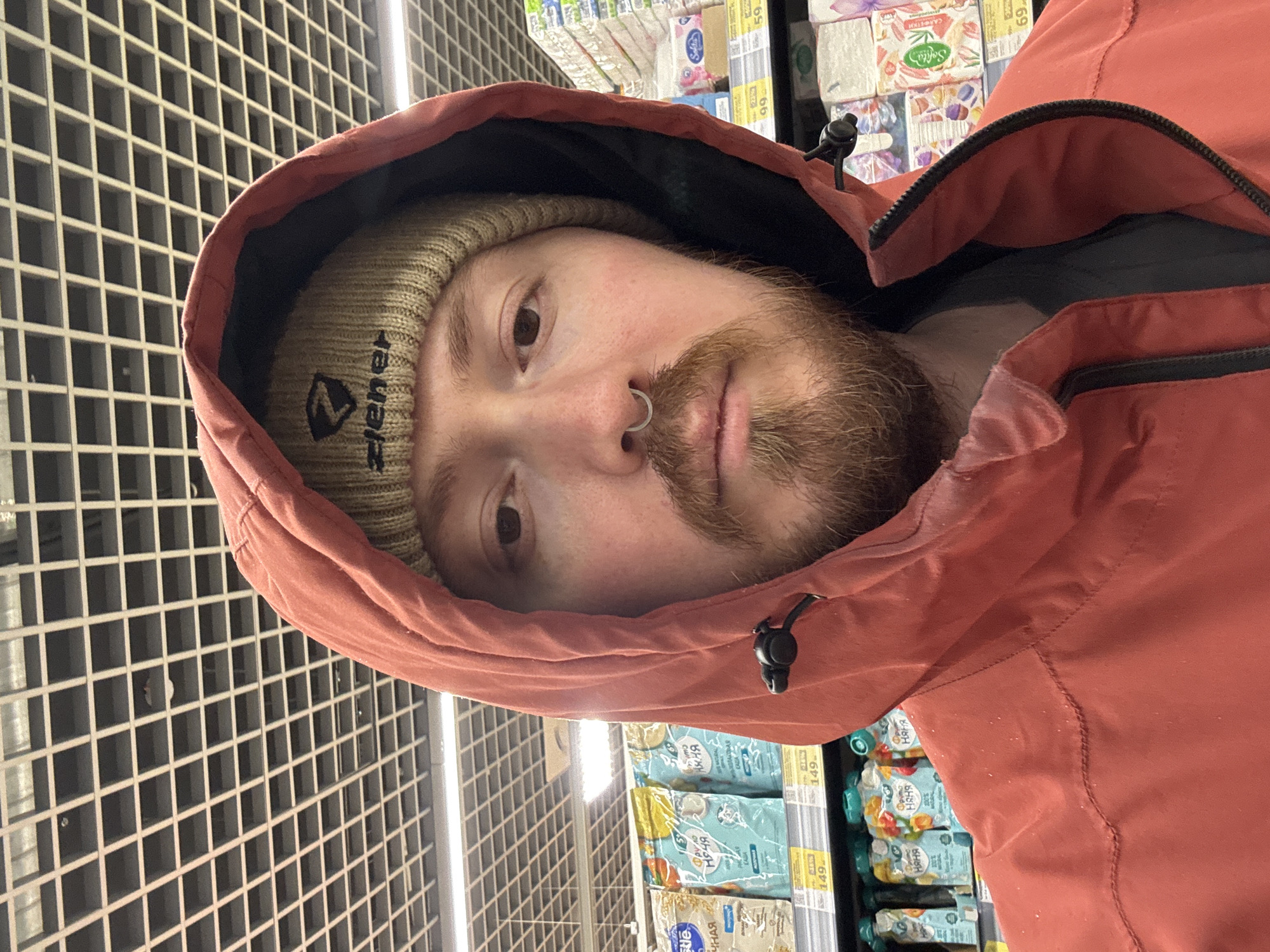 A man with a beard, wearing a beige beanie and a red hooded jacket, is taking a selfie indoors, possibly in a store. He has a nose ring and is looking at the camera. The background shows shelves with various packaged goods and a grid-like pattern above. Some of the items on the shelves have Russian text, and price tags are visible. The text "ЧНАЯ Няня" is legible on the packages. The beanie has the text "ziener" on it. The price tag displays "149". The text "Sofita" is displayed above. The text "САЛФЕ" "59" is visible. The location could be in the city of Chaikovsky, Russia, based on the presence of Russian text on the items in the background.