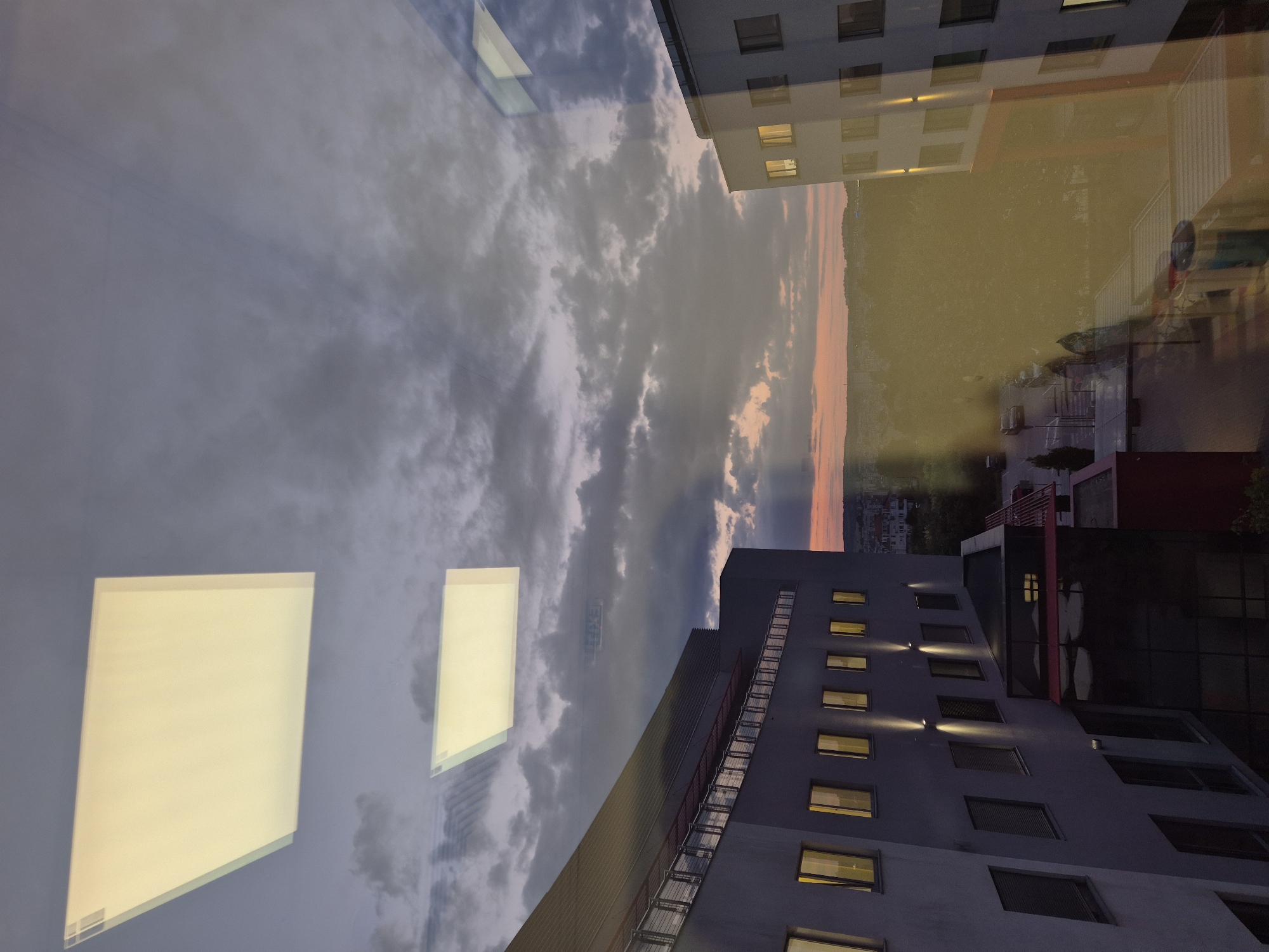 This image captures an exterior view, likely from an elevated vantage point such as a window or balcony, in an urban environment during what appears to be dusk or early evening. The foreground is dominated by reflections on a glass surface, superimposing internal lighting fixtures and structural elements onto the external scene.
On the left, a modern building with a light gray facade is visible. It features multiple windows, some illuminated with a warm yellow light, suggesting interior occupancy. The building has a distinctive sloped roofline and a metal railing system along an elevated walkway or balcony. Spotlights are mounted on the facade, illuminating the wall below.
To the right, another building, primarily white with orange accents, is partially visible. It also has illuminated windows. The reflection on the glass in front of this building shows the same interior lighting fixtures as seen in the reflections on the left side of the image.
The background reveals a city skyline with buildings and trees, bathed in the soft hues of sunset, with shades of orange and pink against a twilight sky filled with dark, dramatic clouds. The overall impression is one of an urban landscape transitioning from day to night, with an emphasis on architectural forms and atmospheric lighting.