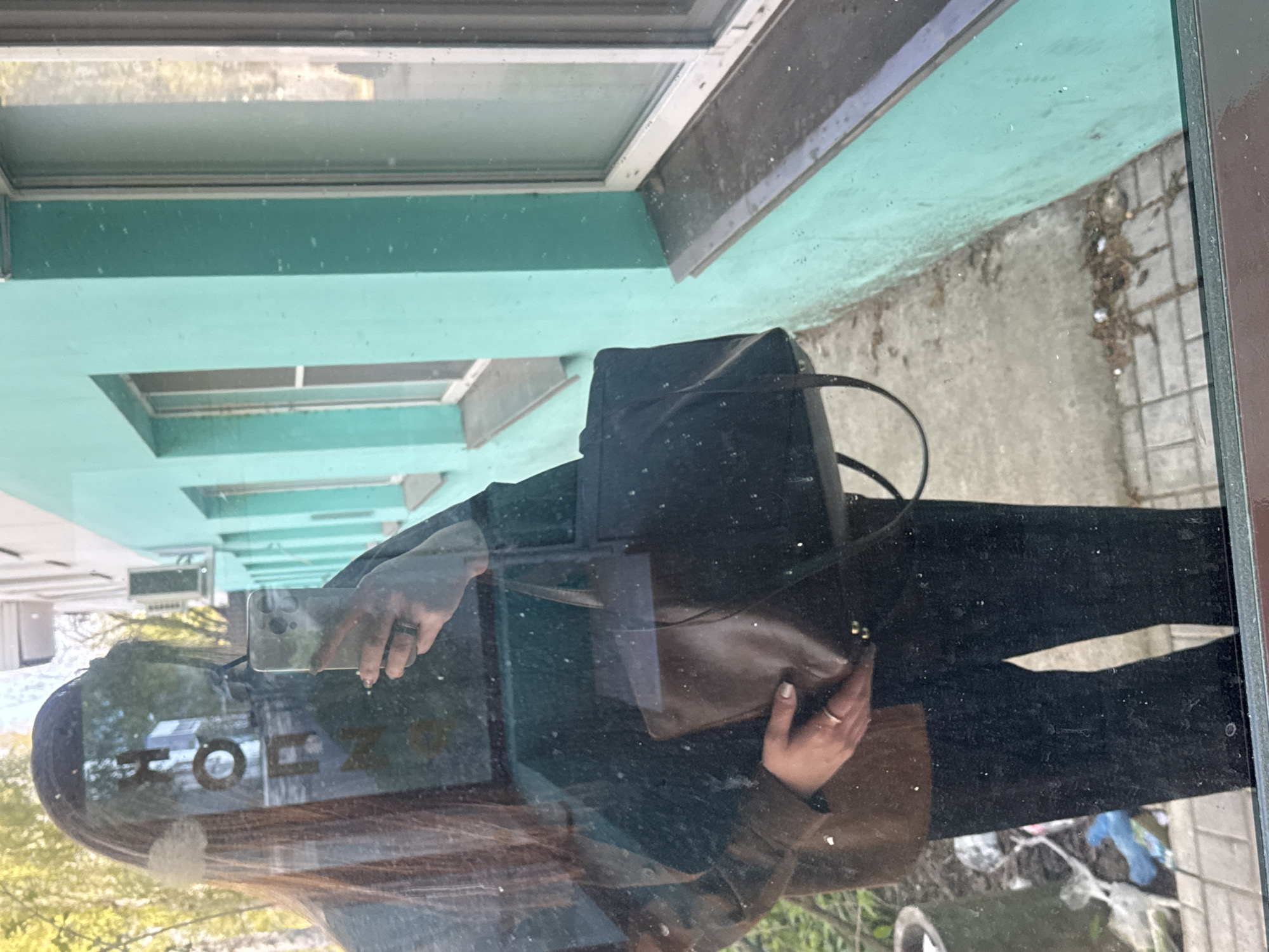 The image shows a reflection in a window. The reflection includes a person holding a phone, presumably taking the photo. The reflection also shows the person's dark clothing and a bag. The reflection is superimposed on the outside view, which includes a green building with window frames and a sidewalk with debris. The reflection also includes the word "TOHZ," reversed. The location is Chaikovsky, Russia.