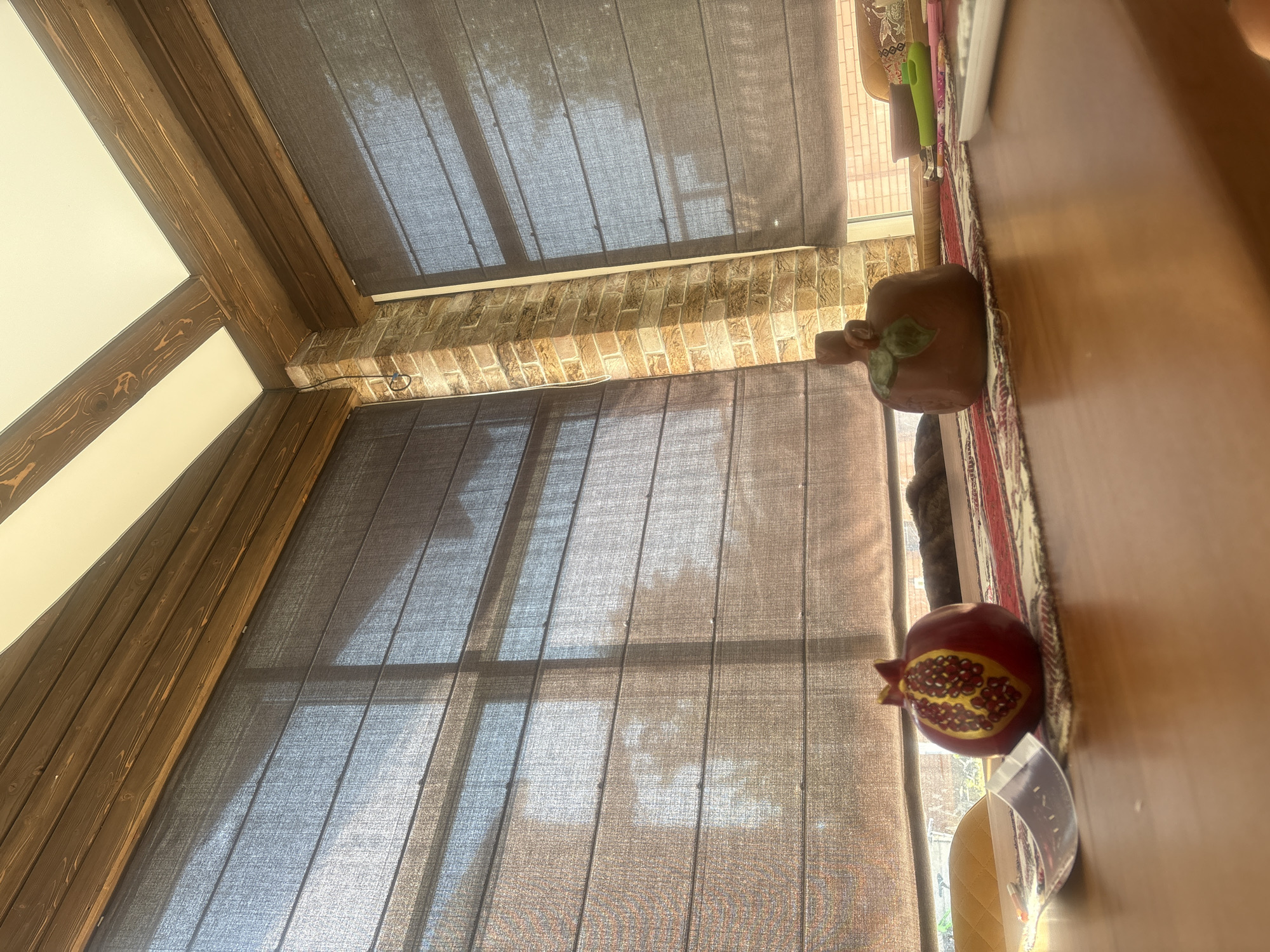 A close-up, low-angle, outdoor shot shows a wooden table with two decorative items, a pomegranate and a pumpkin. The pomegranate is red with yellow and red seed details. The pumpkin is brown with a green leaf detail. Both items are resting on a patterned runner. In the background, large windows with dark roman shades are visible, with sunlight filtering through and casting shadows. The architecture suggests a sunroom or conservatory. The overall setting is bright and airy. The specific location in Georgiyevsk, Russia is unknown.